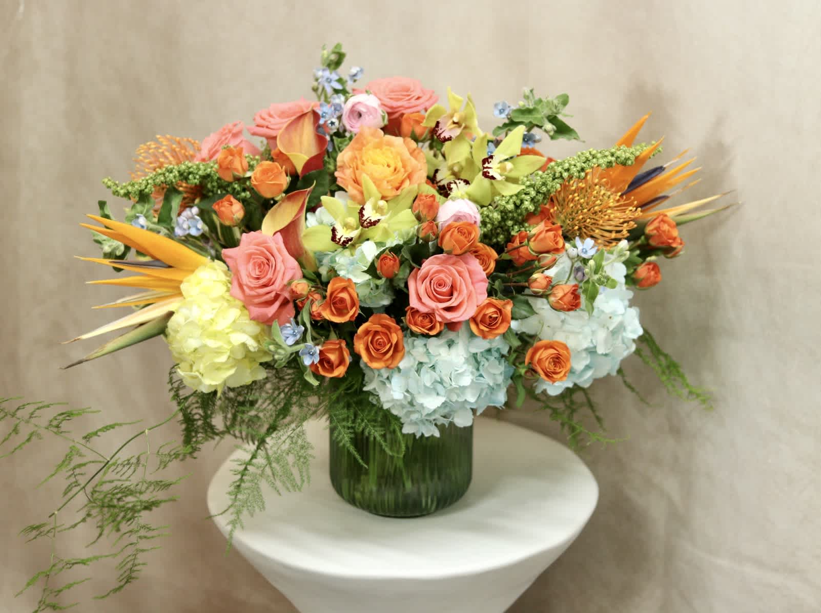 Vibrant Haven  - Introducing &quot;Vibrant Haven&quot; by Venetian Flowers, a stunning bouquet that captures the essence of a tropical paradise. This exquisite arrangement showcases a harmonious blend of vibrant colors and captivating textures, designed to bring a burst of joy and energy into any space.  At the heart of this bouquet, you'll find magnificent green cymbidium orchids, their elegant petals adding a touch of sophistication and grace. Surrounding them are striking blue and greenish yellow hydrangeas, creating a captivating contrast that mimics the vibrant hues of a tropical landscape.  Adding a touch of exotic allure are the majestic birds of paradise, with their vivid orange and blue hues resembling the fiery plumage of tropical birds. These extraordinary blooms truly make a statement, commanding attention with their unique shape and vibrant colors.  Nestled among the birds of paradise are the radiant orange free spirit roses, orange spray roses, and coral pink roses. These blooms exude warmth and passion, symbolizing the energy and enthusiasm found in tropical destinations. Their delicate petals and sweet fragrance add a romantic touch to the arrangement.  Accentuating the bouquet are the cheerful orange mini calla lilies, the delicate blue tweedia flowers, and the eye-catching orange pin cushion protea. These elements provide an extra layer of depth and visual interest, creating a multi-dimensional arrangement that is as captivating as it is vibrant.  To complete the presentation, &quot;Vibrant Haven&quot; is artfully arranged in a modern vase made of blue cut glass. The sleek design of the vase adds a contemporary touch, perfectly complementing the bold and tropical aesthetic of the bouquet.  Whether you're looking to brighten up your home, surprise a loved one, or add a touch of tropical elegance to a special occasion, &quot;Vibrant Haven&quot; by Venetian Flowers is the perfect choice. It brings the beauty and vibrancy of a tropical paradise right to your doorstep.