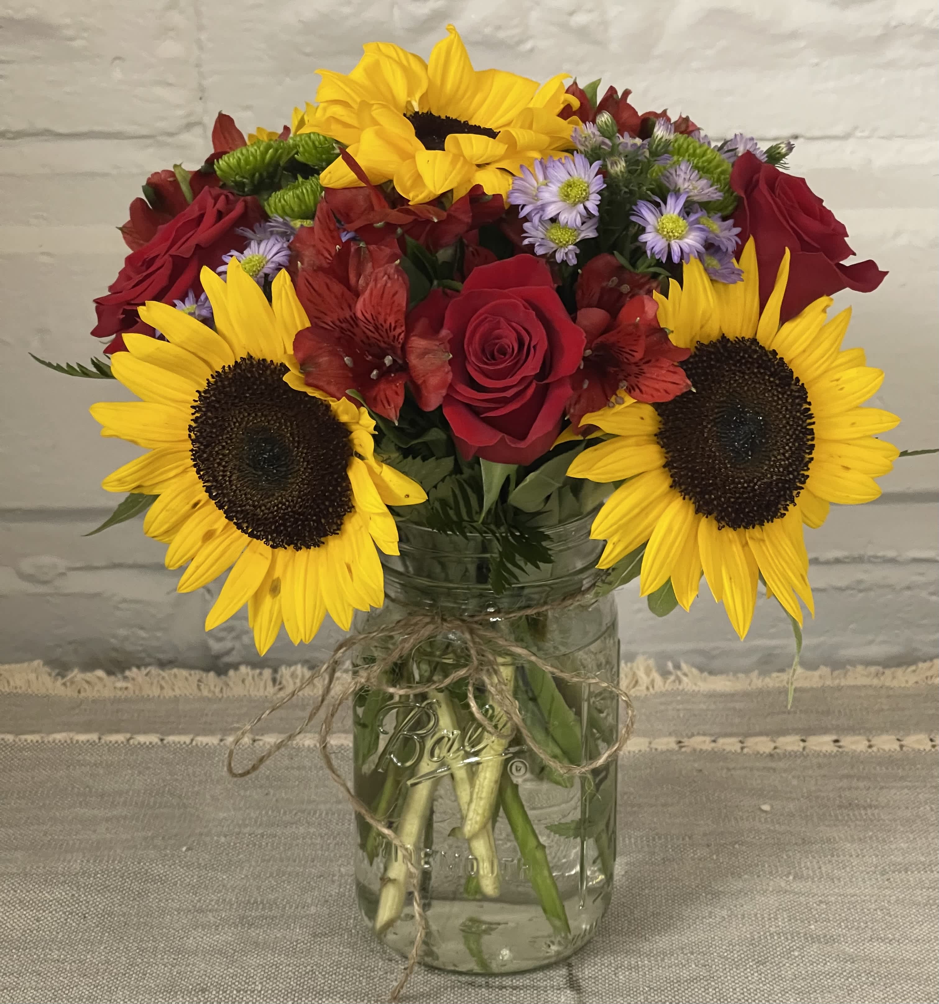 Janie's Sunshine Bouquet - This delightful arrangement that captures the essence of a sunny day in the countryside. This charming bouquet features vibrant sunflowers, symbolizing happiness and positivity, paired with elegant roses for a touch of romance. Nestled in a classic Mason jar, adorned with a rustic string tied around it for that extra country-style flair, this arrangement is perfect for adding warmth and cheer to any space.
