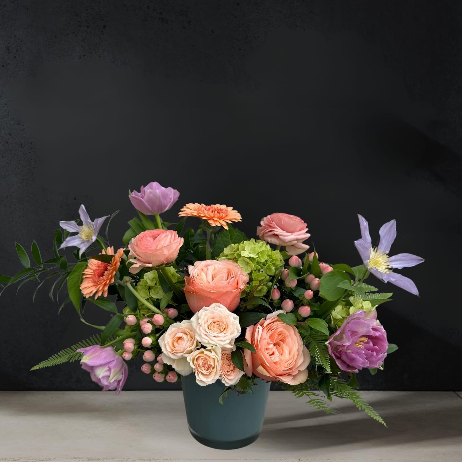 Coral Charm - A sleek gray glass vase cradles a one-sided arrangement of soft peach blooms, elegantly accented with lush green foliage and delicate lavender flowers