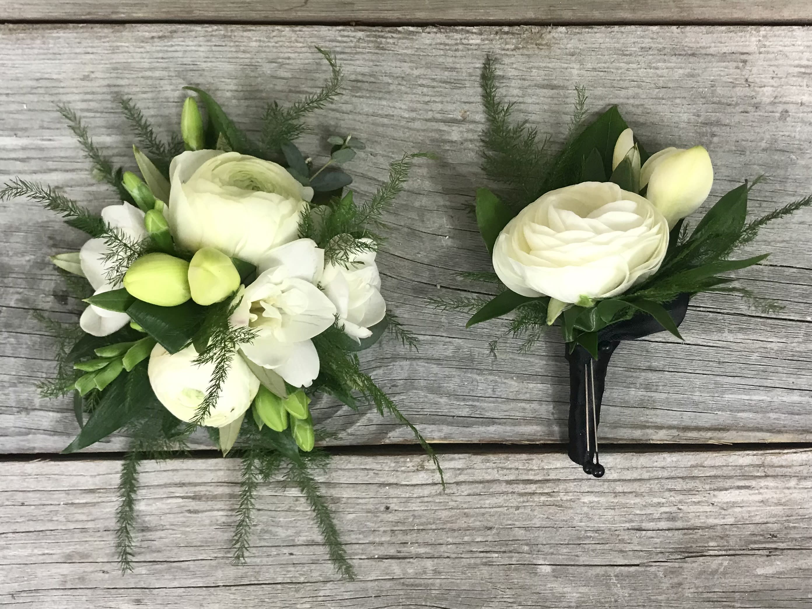Free Spirit Corsage Set - A wrist corsage and matching boutonniere of white ranunculus, white freeisa accented with greenery. Substitutions may be made if needed.