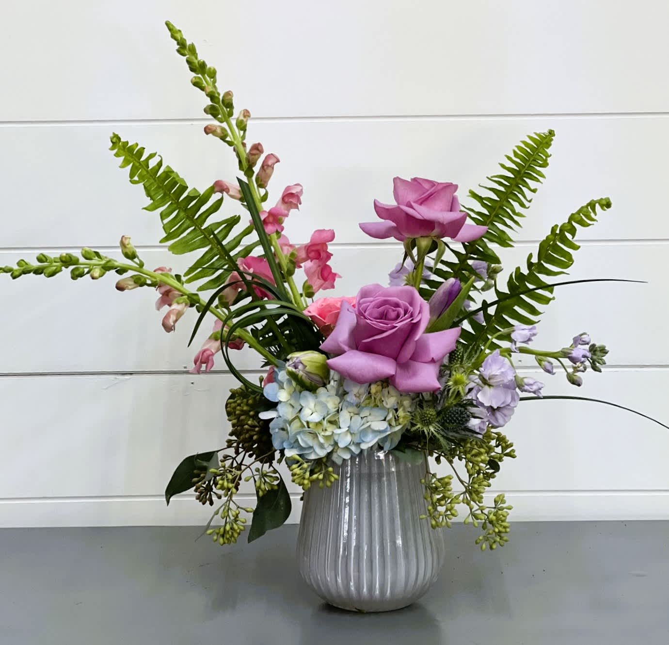 Blooming - This lovely lavender keepsake vase is sure to be a hit on your Easter or Spring table. It is filled with lavender reflexed roses, blue hydrangea, tulips, spring greenery and filler.