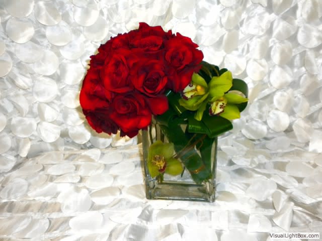 Custom Arrangement with Roses and Cymbidium Orchid - Custom arrangement with a cluster of roses, ti leaves and cymbidium orchid blossoms outside. Submerged inside a square vase. Dimensions: 14-18 in height, 10-14 in length.