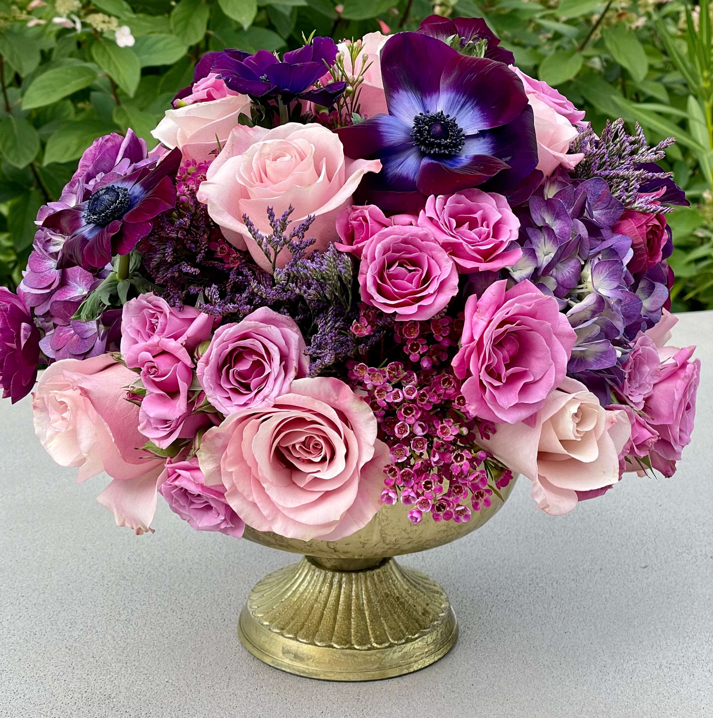 Kika - A blend of purples in light and deep hues in an elevated footed compote. Perfect for a dinner party or event centerpiece. Deep Anemones and Pale purple roses are the showstoppers here!