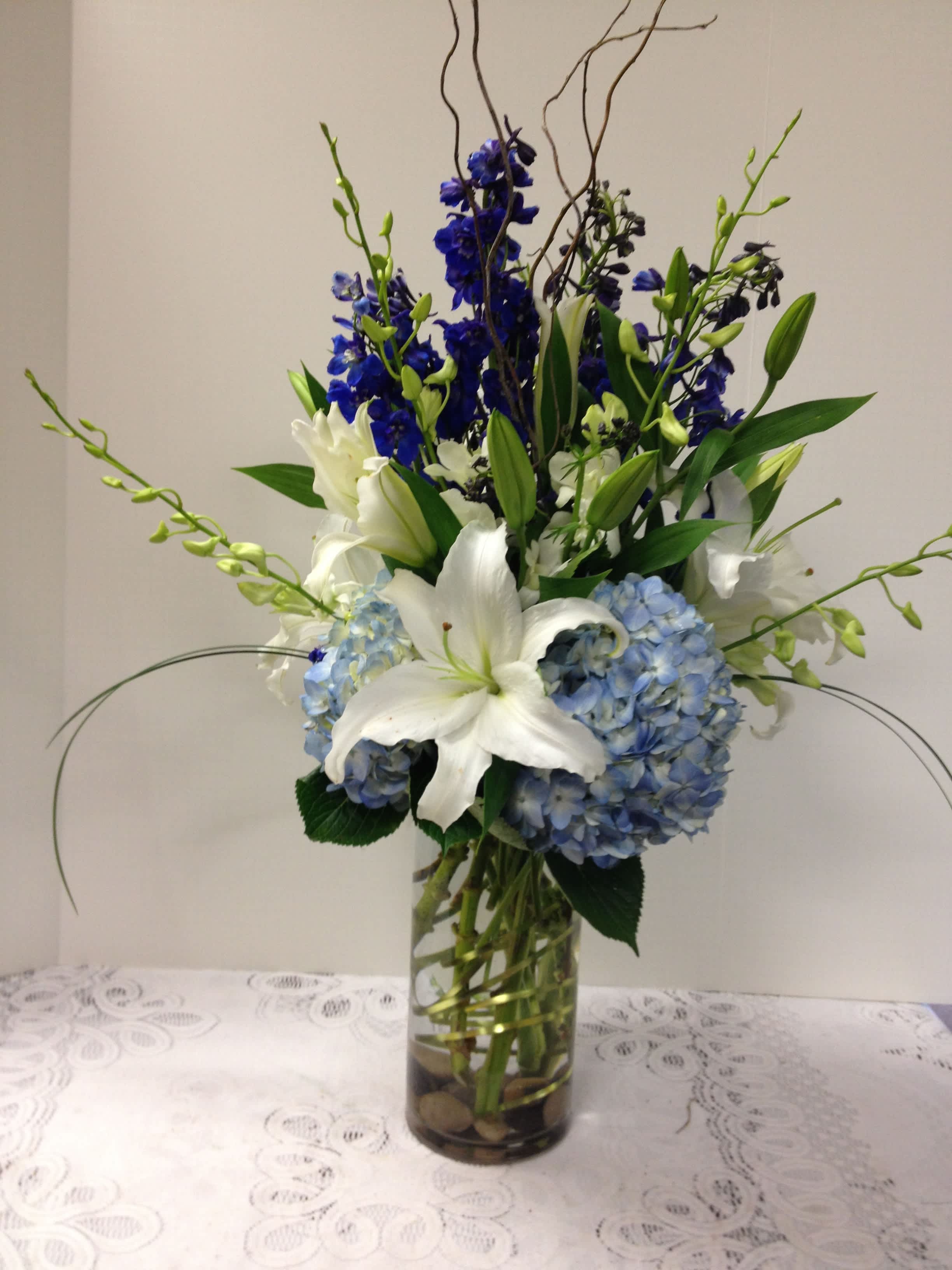 Classy - A clear glass container filled with southern blue hydrangea, fragrant white lilies, dendrobium orchids and showy curly willow accented with lily grass.