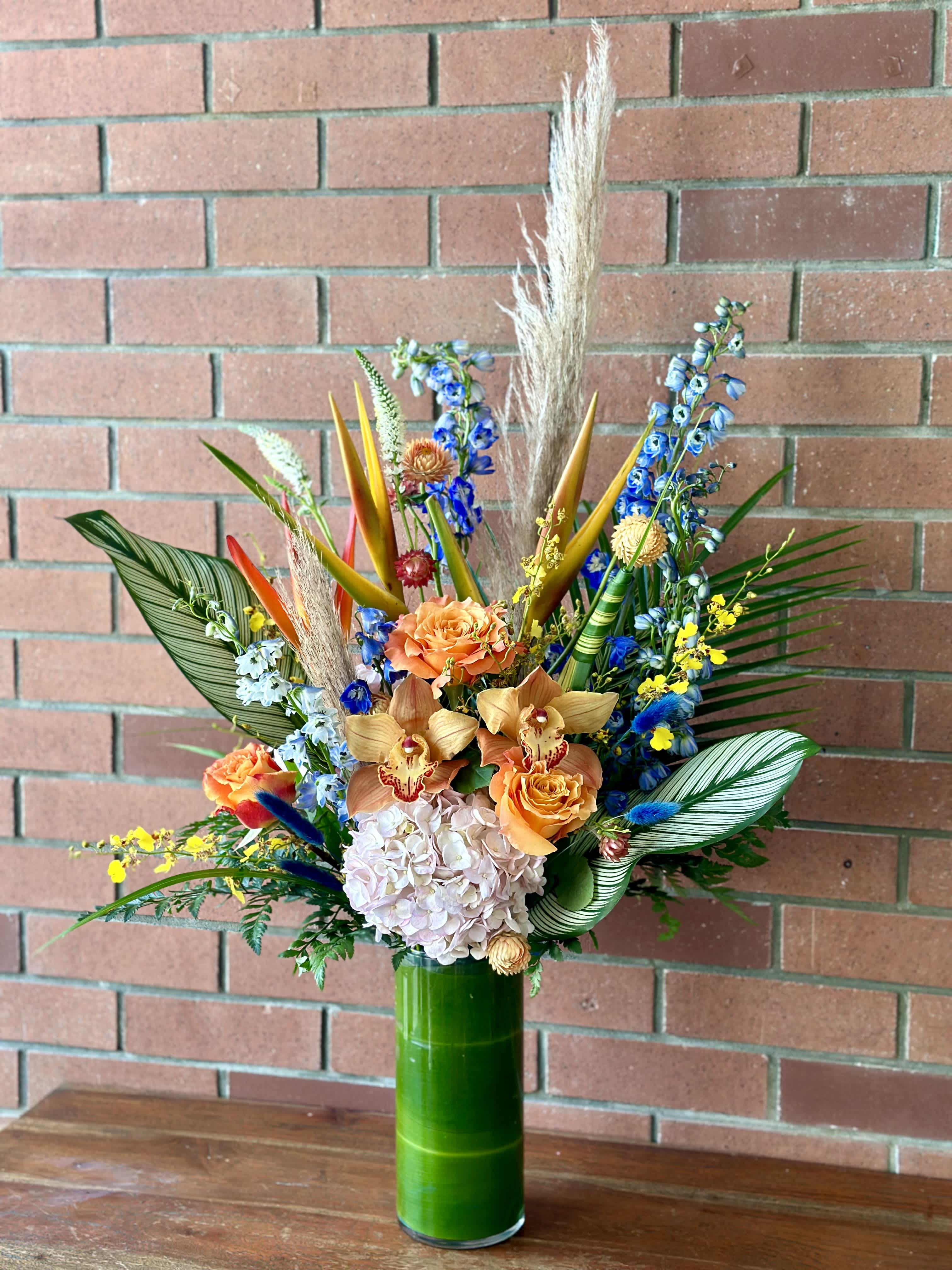 Floridian Beauty Bouquet  - Send a vibrant elegant / tropical bouquet of blooming delphiniums, orchids, free spirit roses, tropical flowers, pampas grass and more. Arranged in a cylinder vase wrapped with green leafs 