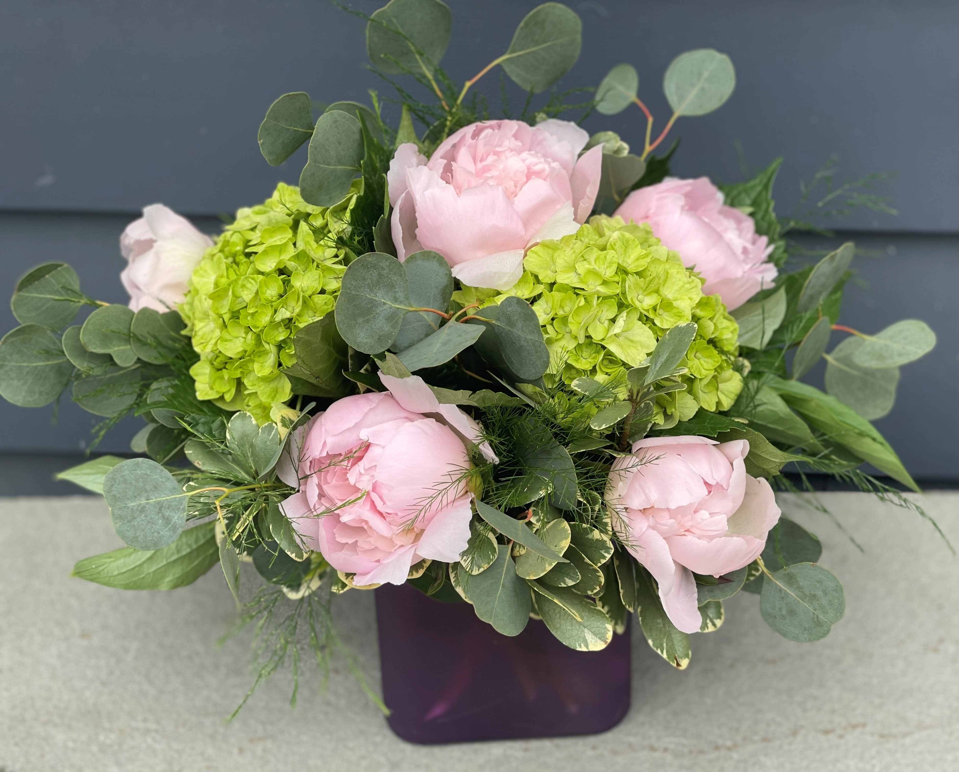 Petite Peony - These pretty pink peonies and their sweet scent are a fan favorite. Arranged in a lavender square vase, accompanied by lime green hydrangea, eucalyptus and other foliage's. *** Shade of peony and vase may vary.