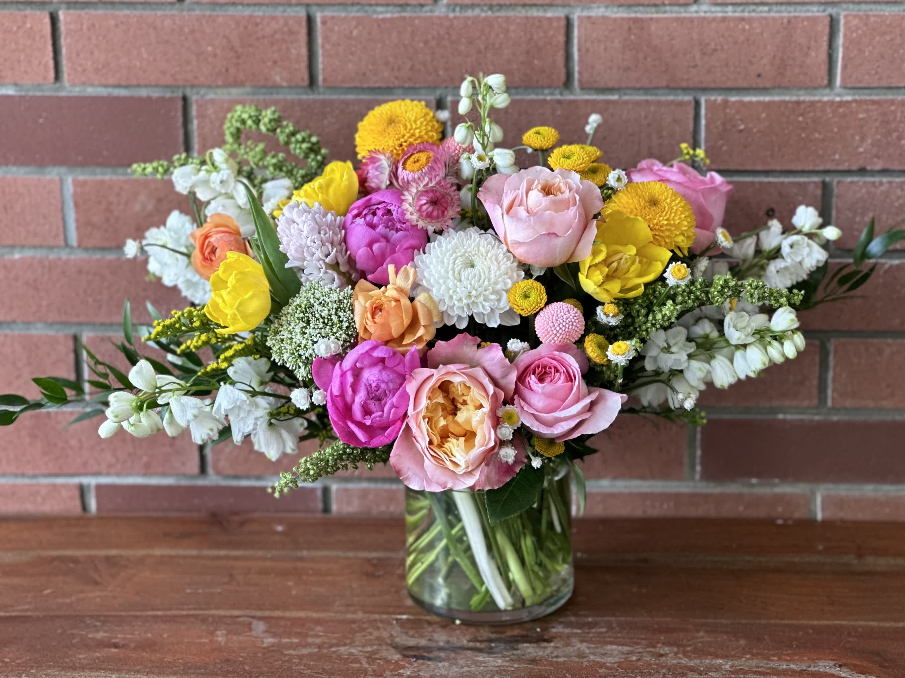 Sunshine Garden  - A spring arrangement featuring peonies, yellow tulips, white mums, straw flowers, white delphinium, garden roses, and ball mums is a beautiful combination of colors and textures.  The combination of these vibrant spring flowers, with their varying colors and textures, creates a visually stunning and dynamic bouquet that is sure to make a statement and bring joy to any space. 