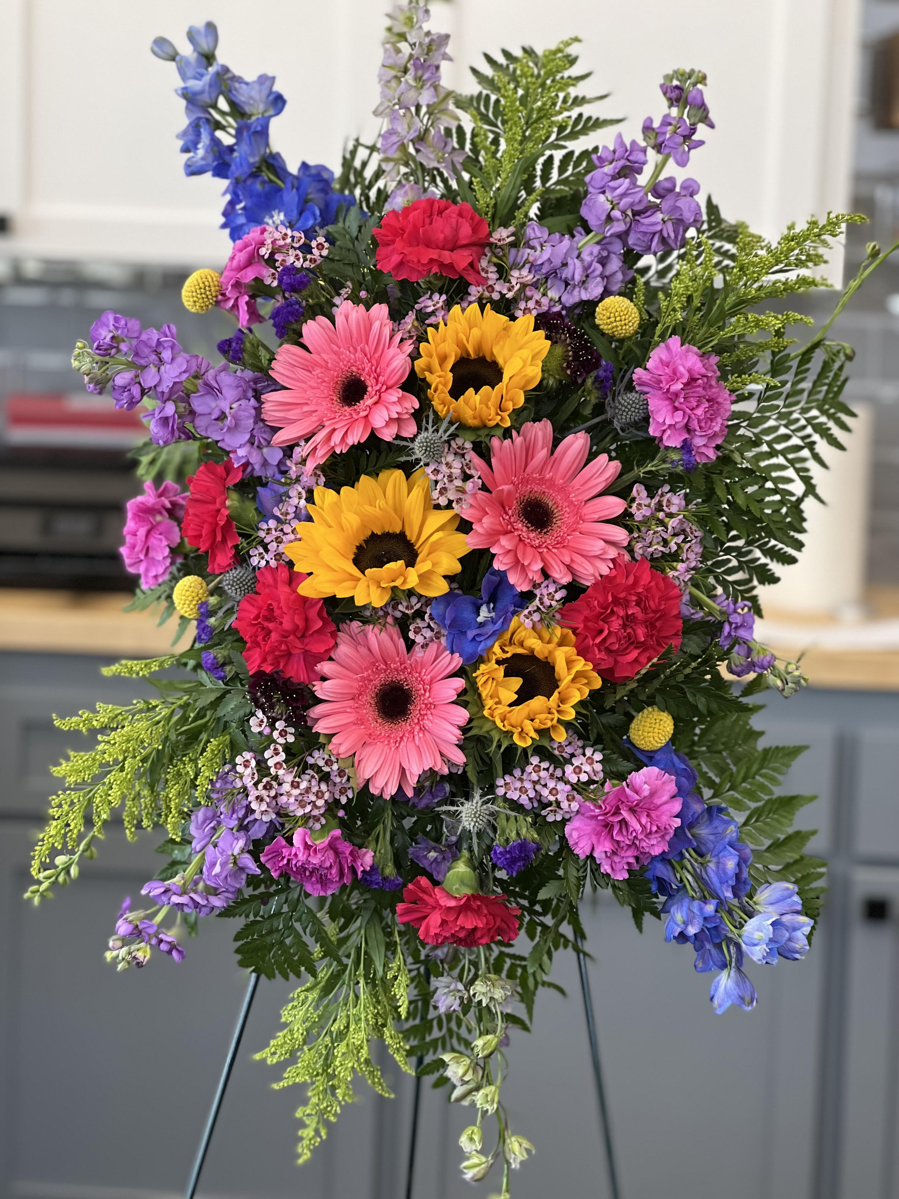 Love Lives On Standing Spray by Anderson Florist - . This spray features hot pink gerberas, hot pink and light pink carnations, medium yellow sunflowers, blue delphinium, solidago, cresipedia, lavender stock, wax flower, blue thistle  and mixed greenery. Wire Easel Needs to be ordered separately. 