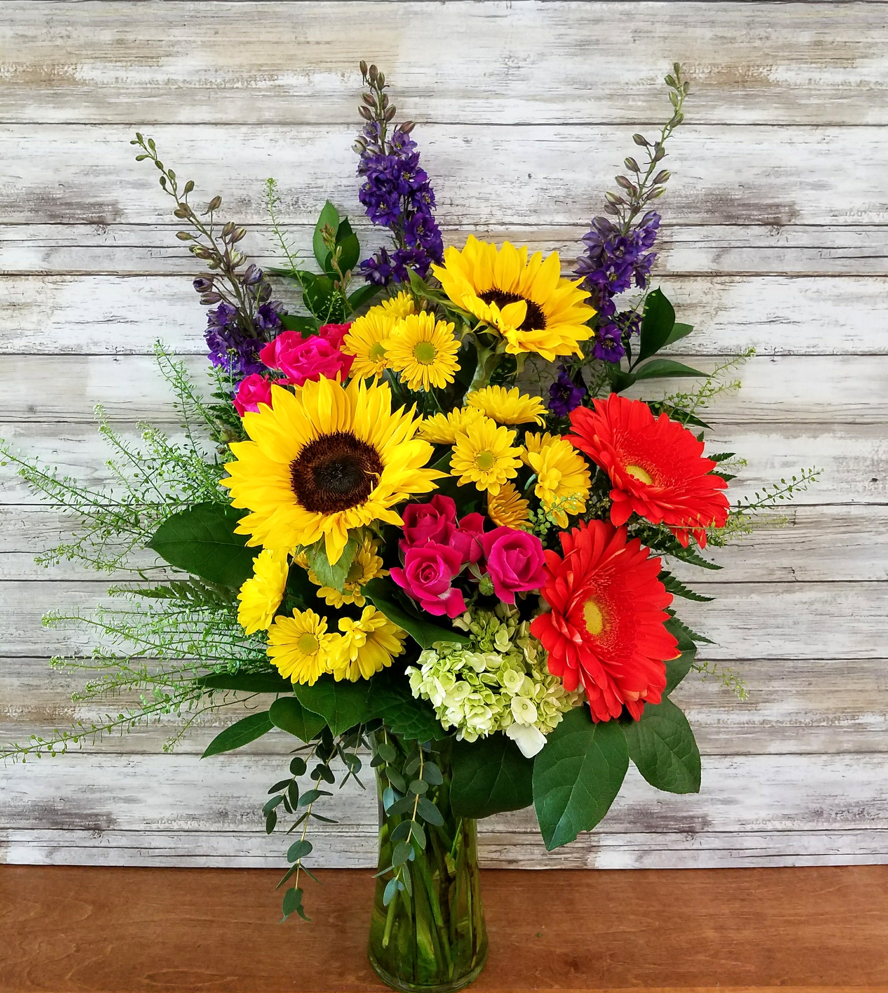 Summer Fiesta - Bright bold colors will surely make them smile!  Gerbera daisies, sunflowers, hydrangea, spray roses, daisies and larkspur arranged with various greens.  Approximately 24&quot; tall 
