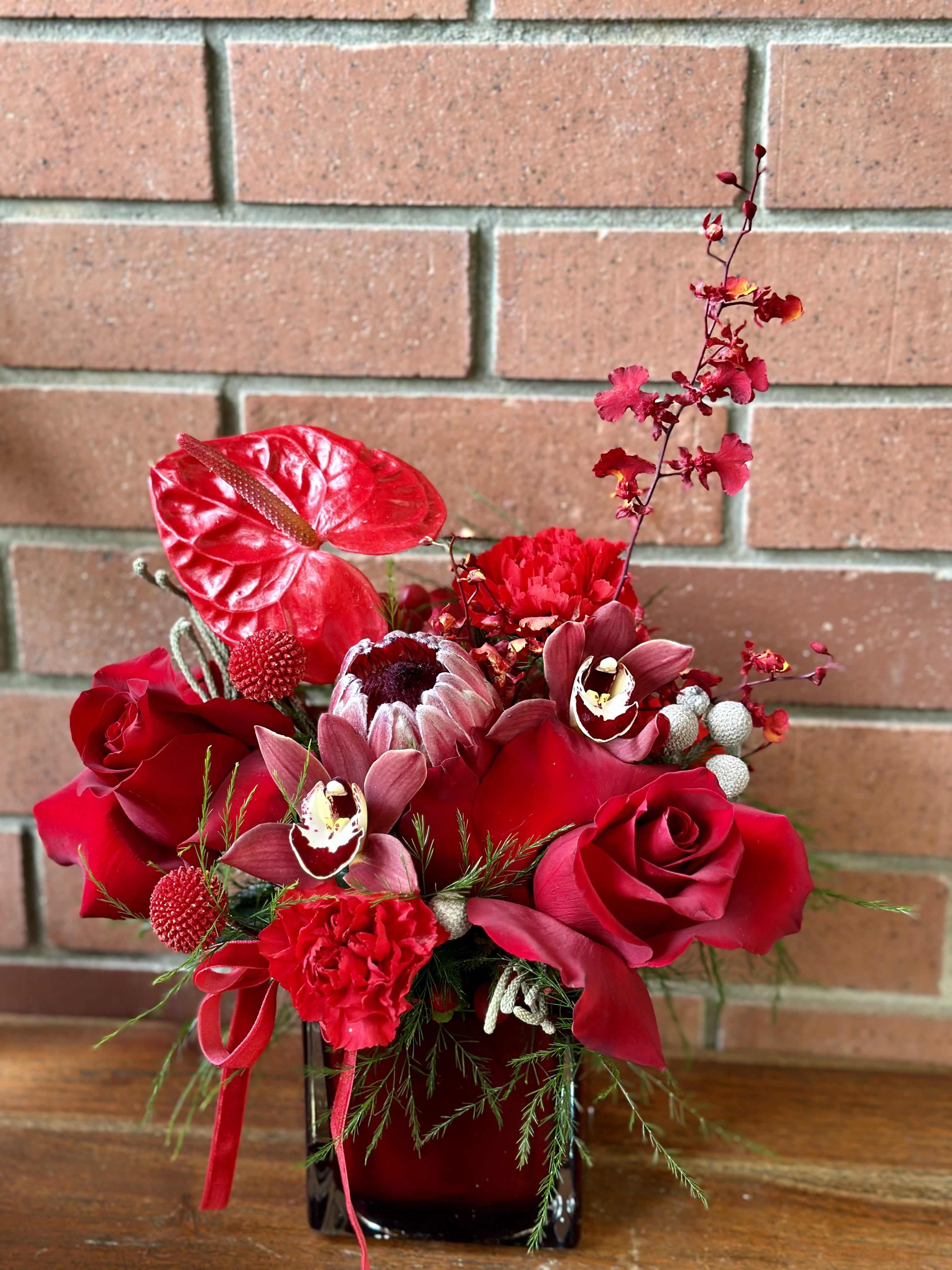 Love Always Bouquet  - Modern ruby red square vase premium flowers - red roses, red orchids, red proteas, red anthurium, touches of red carnations and red craspedia - premium greenery and fillers accented with a velvet red ribbon. Tropical elegant bouquet. Deluxe and premium are designed in a 5x5 red square ruby vase. 