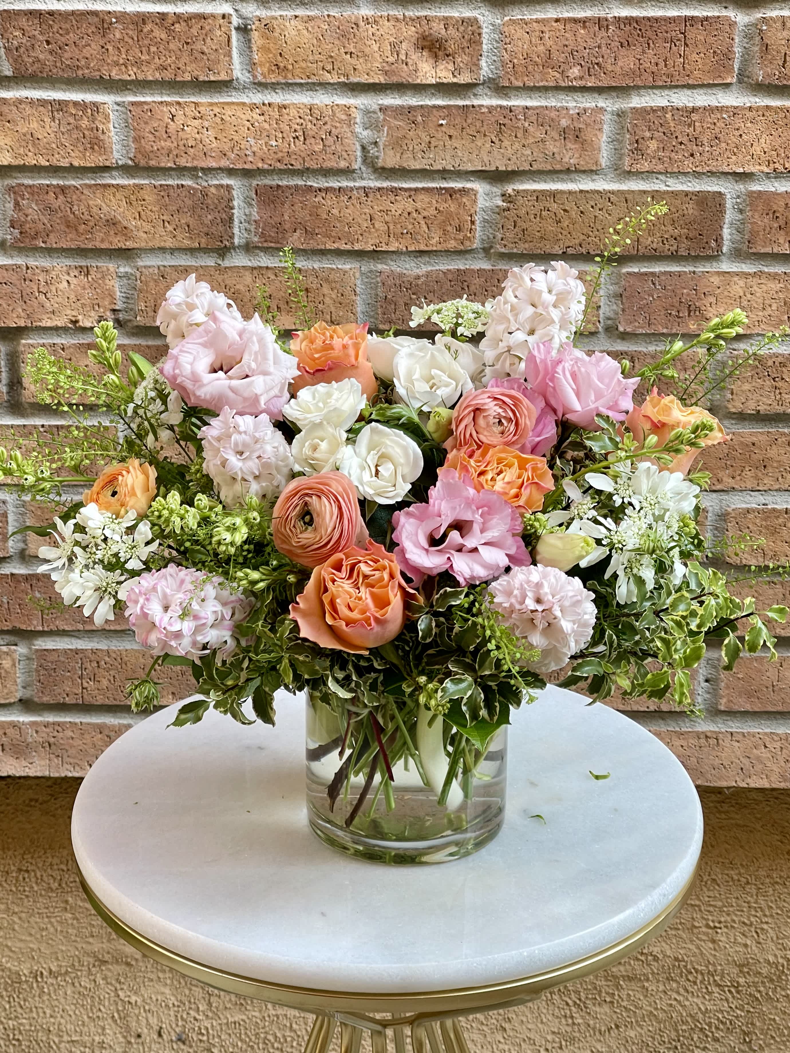 Featured Design - Every week we feature a new design that includes some of our favorite flowers. Available for a limited time only.  This arrangement of beautiful shades of pinks, coral and ivory flowers in a clear glass cylinder vase includes hyacinth, roses, ranunculus, lisianthus, majolica spray rose, variegated Italian pittosporum and thlaspi foliage.  Vase dimension: 5”W X 5”H