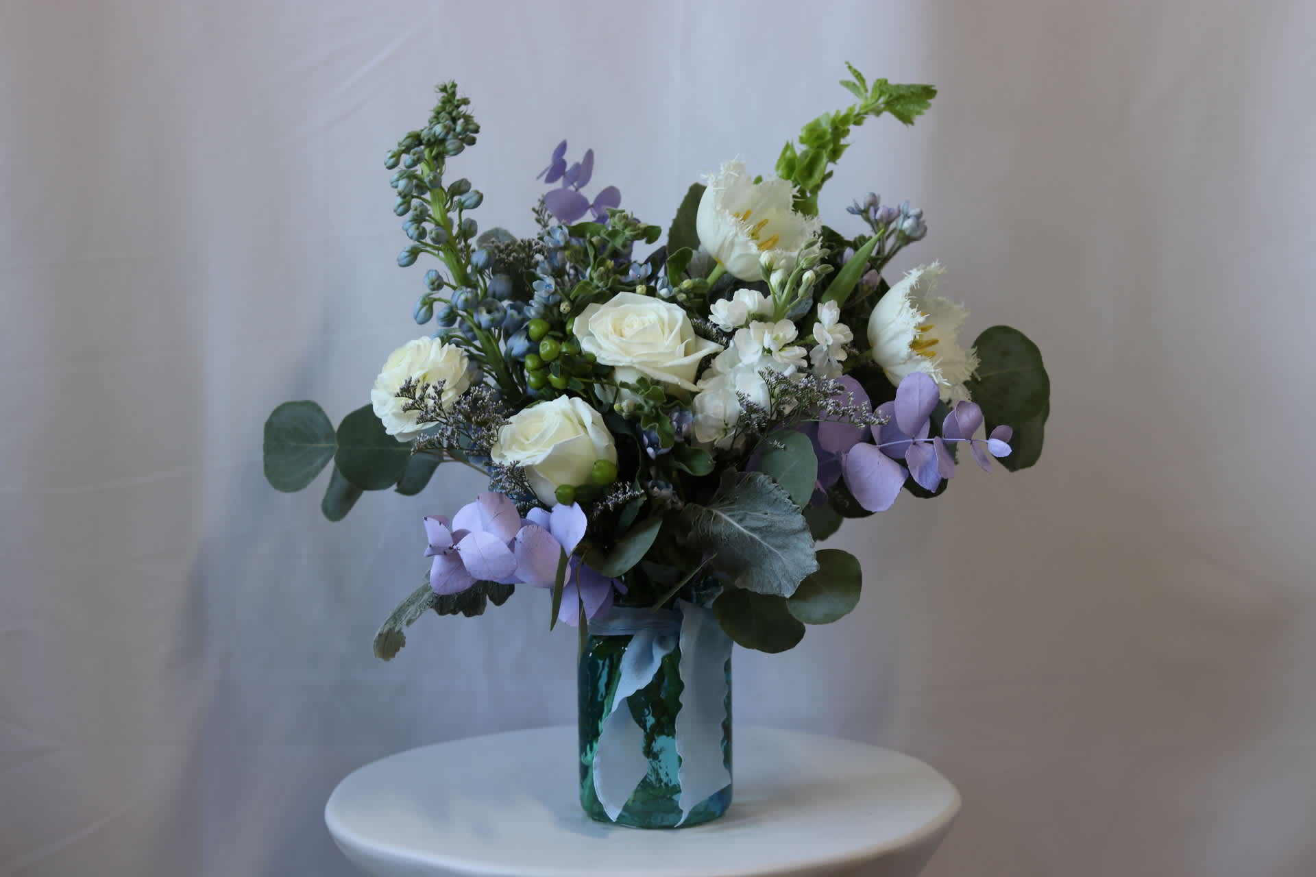 Skyfall Serenity Bouquet  - The Venetian flower bouquet is a stunning arrangement that showcases the beauty of various flowers in a 5&quot; blue hammered jar. The bouquet features a combination of blue delphinium, white roses, and white tulips, which create a harmonious blend of colors. The green bells of Ireland add a touch of freshness and vibrancy to the arrangement.  To enhance the bouquet's elegance, dusty miller and lavender painted eucalyptus are incorporated, providing a delicate and fragrant accent. The bouquet is tied together with a blue ribbon, adding a final touch of sophistication.  With a bouquet size of 12&quot;, the Venetian flower arrangement is sure to make a statement and bring a touch of Venetian charm to any space or occasion.