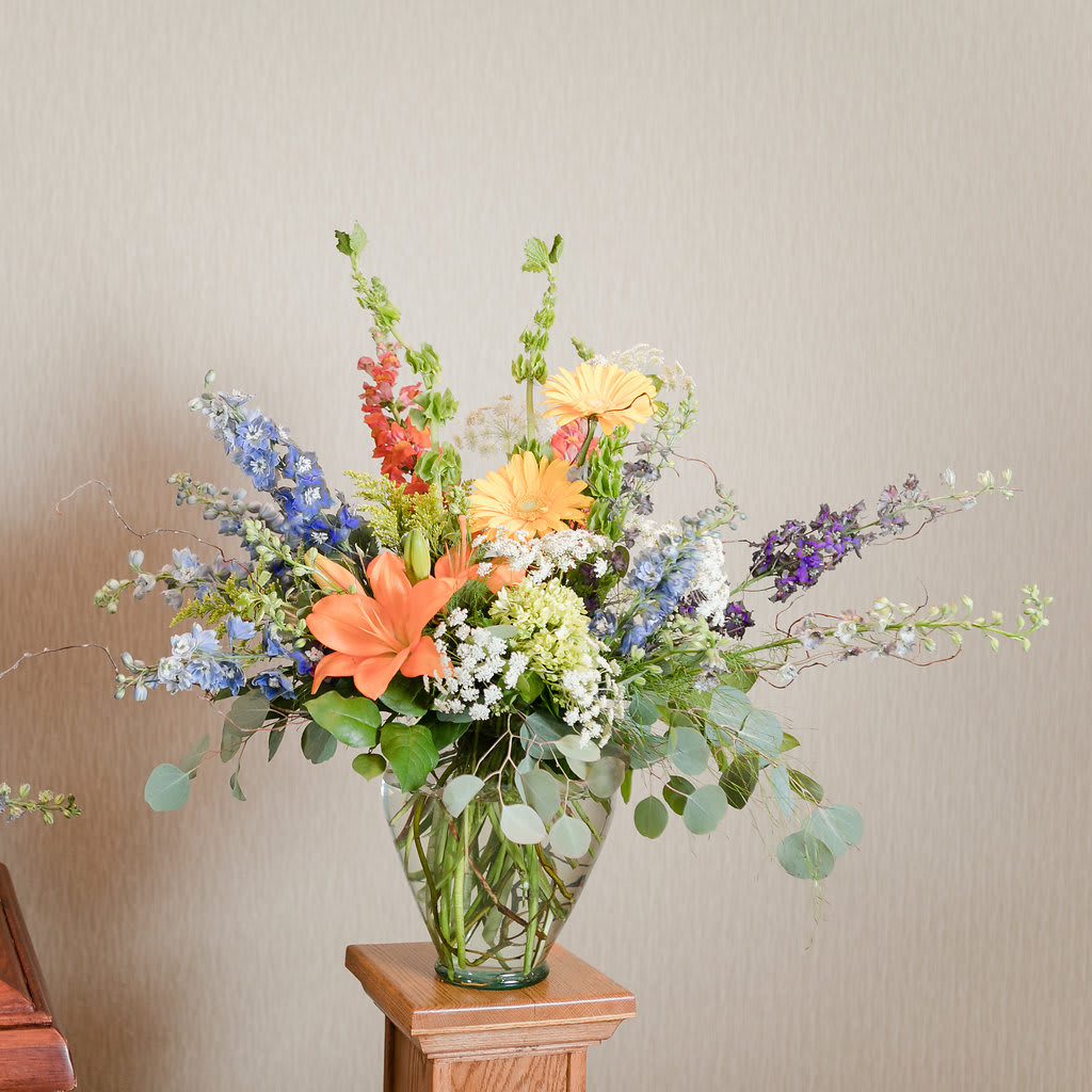 Barefoot - A bright, wildflower like arrangement with orange lilies, blue delphinium and yellow gerbera daisies, perfect to honor a vibrant life. 