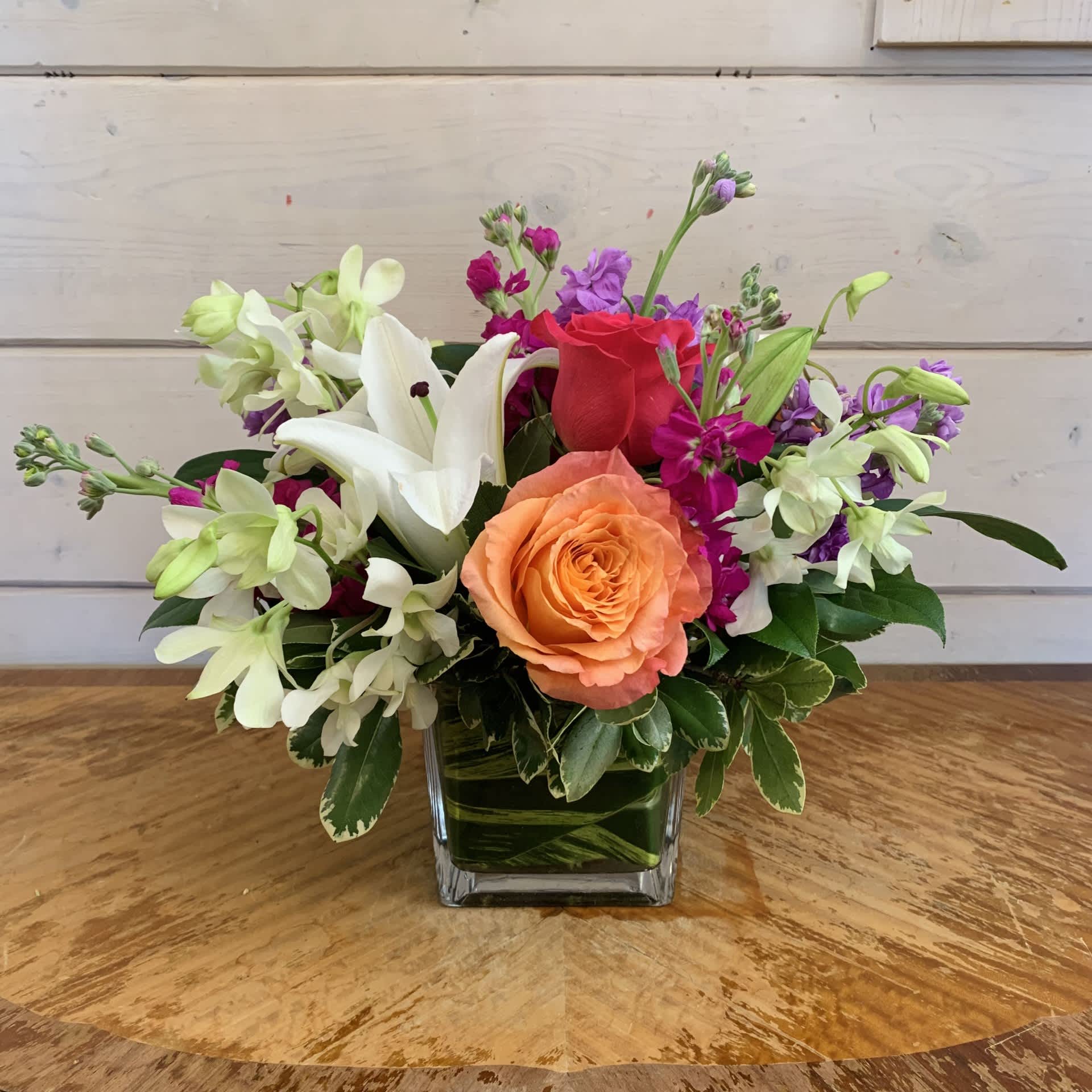 Make Her Day Bouquet  - White orchids, hot pink and orange roses with a white lily arranged in a medium leaf lined cube with magenta and lavender stock. Approximately 10in tall and 14in wide. 