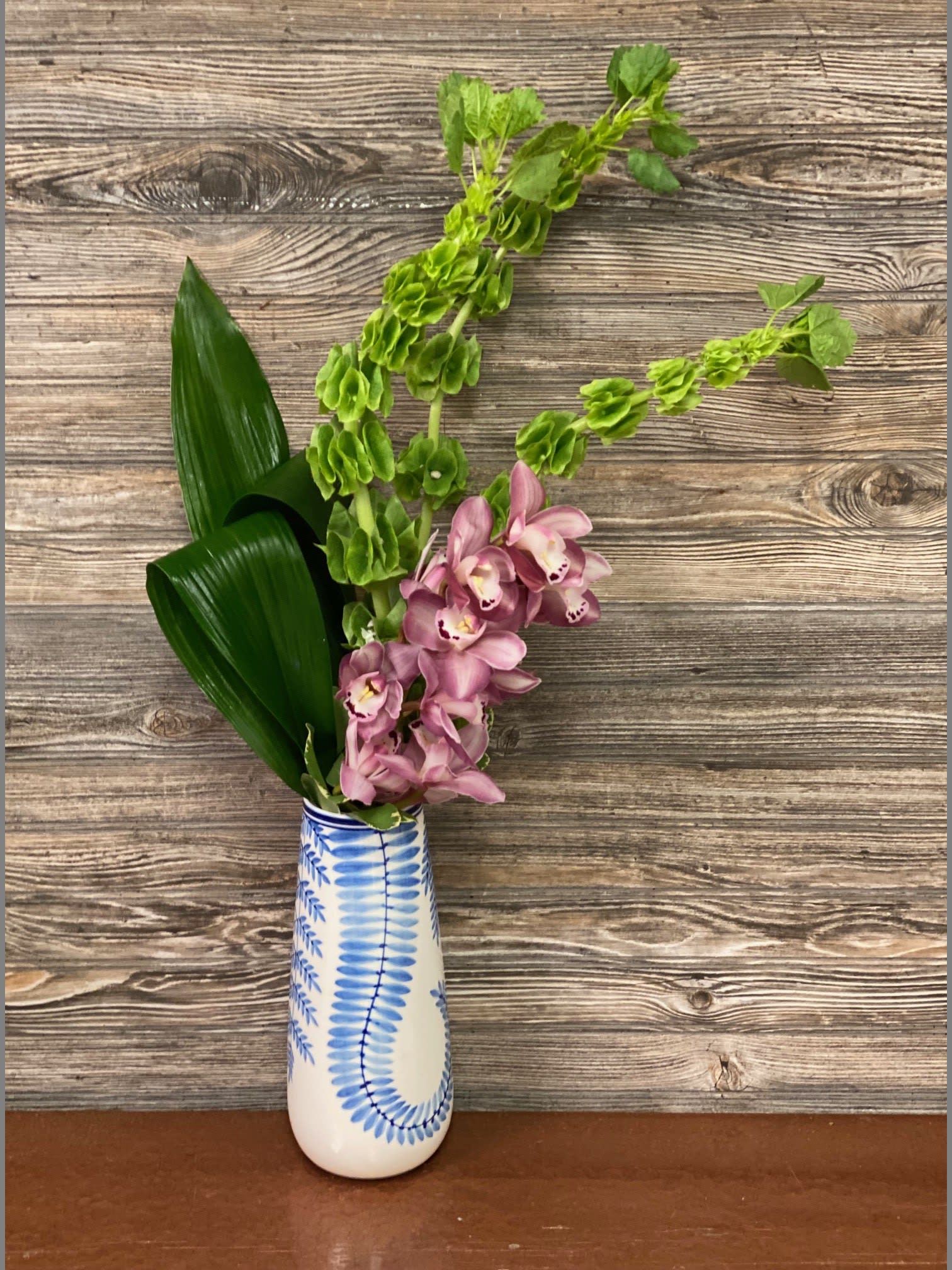 Modern Beauty - Tropical Cymbidium Orchid stems arranged with Bells of Ireland and aspidistra leaves in a Blue Delft vase. 1 stem for Standard, 2 stems for Deluxe, 3 stems for Premium approximate height 27&quot;
