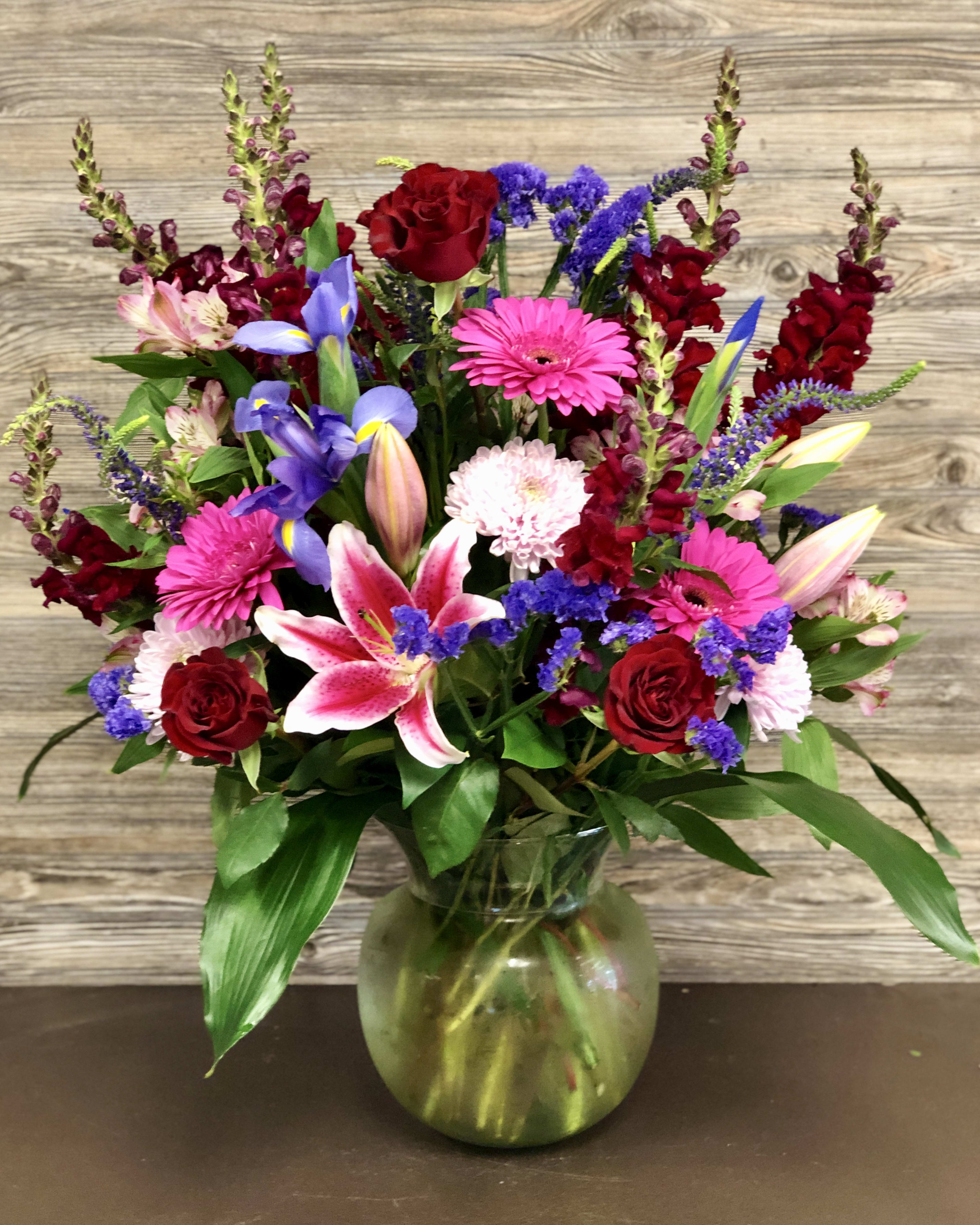 Vibrant Garden Mix  - Assorted mixed seasonal flowers like lilies, roses, gerberas, iris, and snapdragons accented with fillers and greenery in a clear rounded Optic vase (Dimensions of the container are 9.75inHx 7.25inW) Arrangement is about 24hx14w.