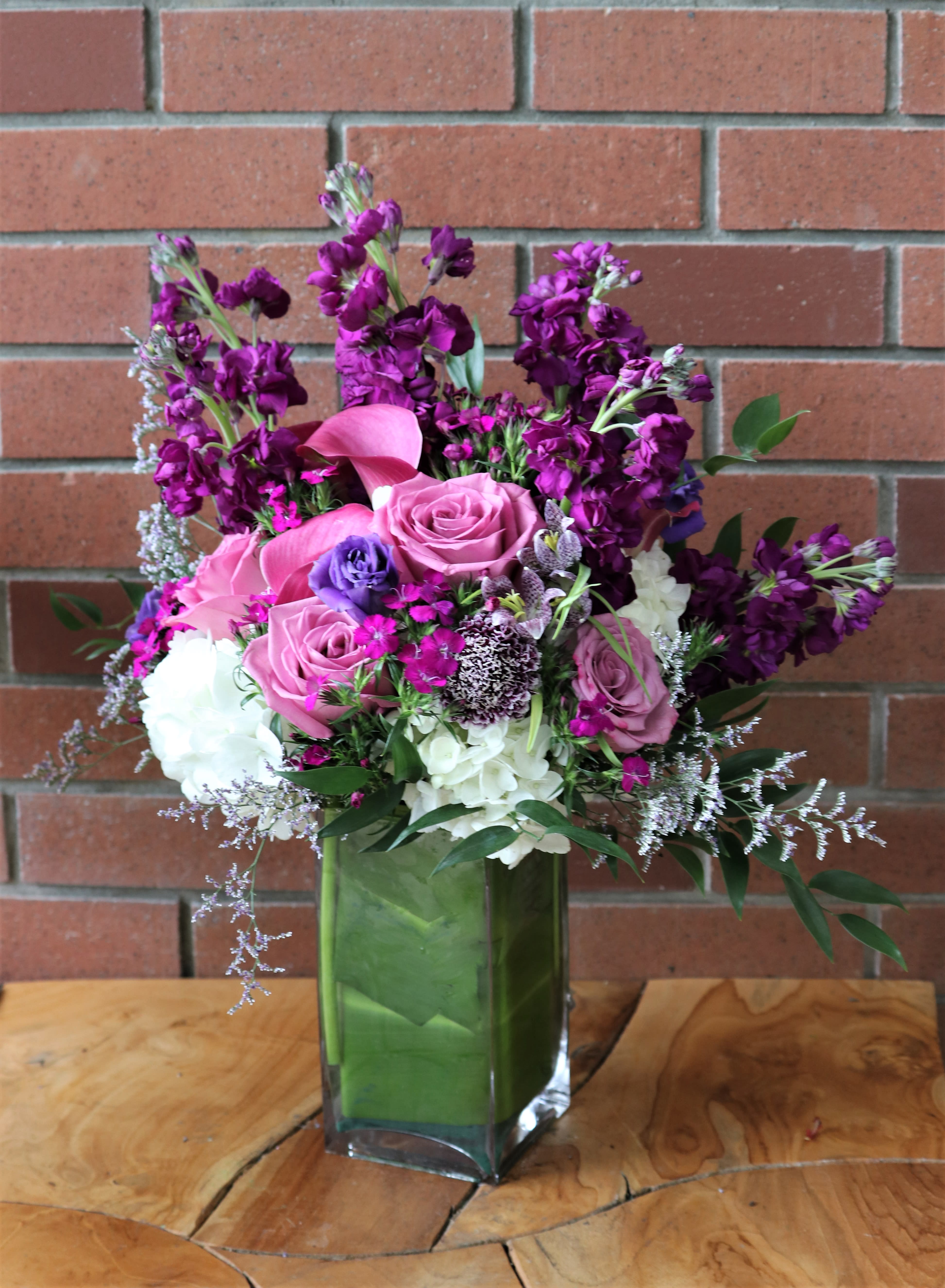 Plum Crazy - Wonderful fresh selection of purple, plum flowers - elegant roses hydrangeas, stock, calla lilies anemones wild flower fillers and more in a modern rectangular vase wrapped with a green leaf. 