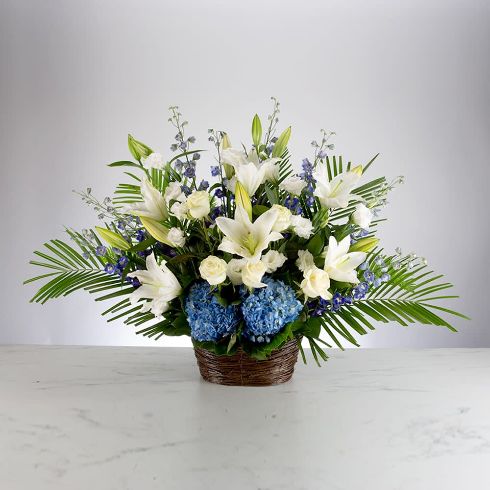 Deepest Condolences - A blue, white, and green funeral basket for any type of service. Held in a wicker basket, large palms spread out and give a natural spotlight for the feature blooms and colors.