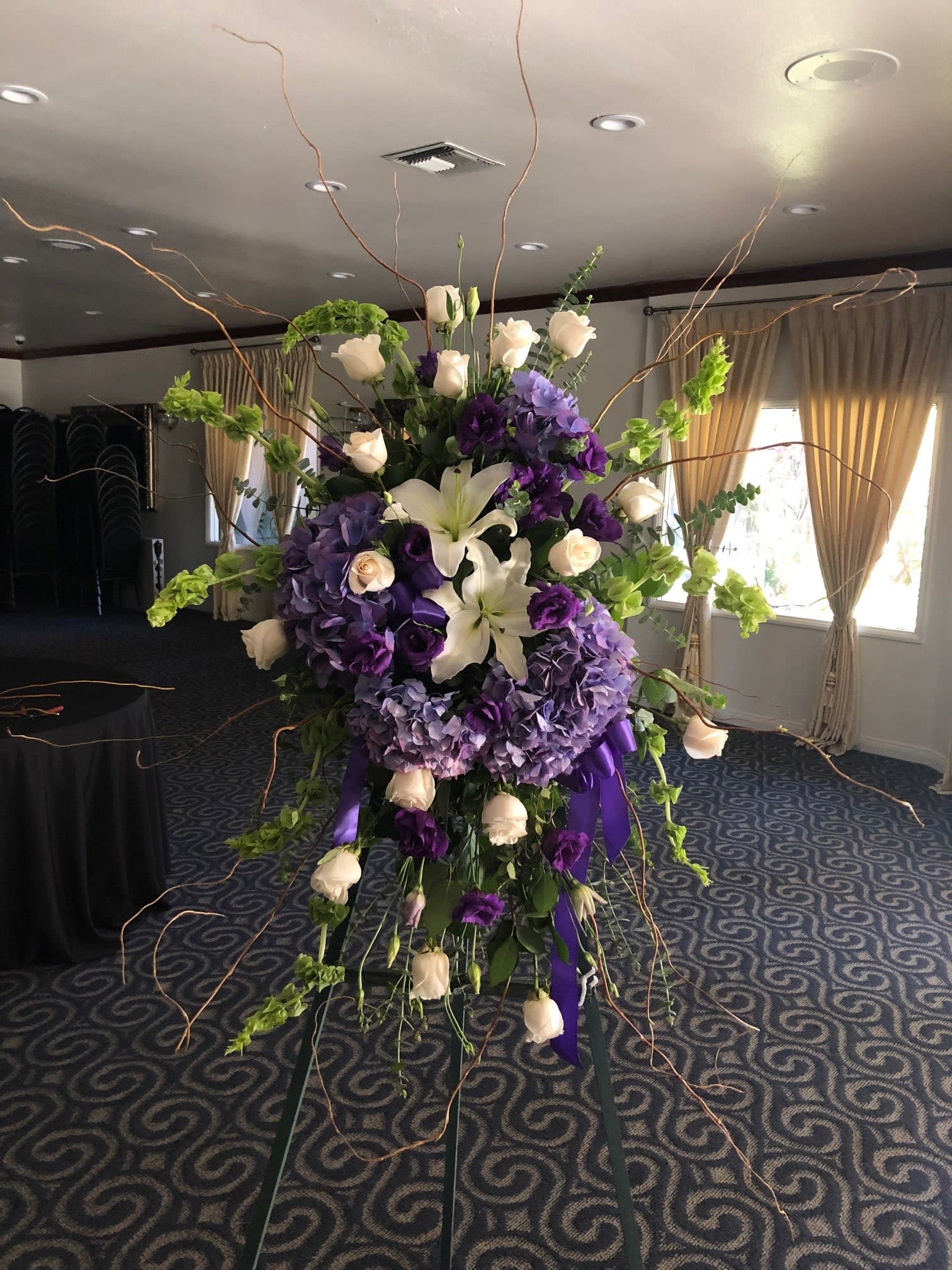 Purple and White Sympathy Spray Piece  - Type of Flowers: White Roses, Purple Hydrangeas, Bells of Ireland, White Asiatic Lilies, White and Purple Lisianthus, Branches, and Ribbon on an easel. Availability: All year round Substitute Available: Yes Design View: All Facing View Photo shown: Regular 