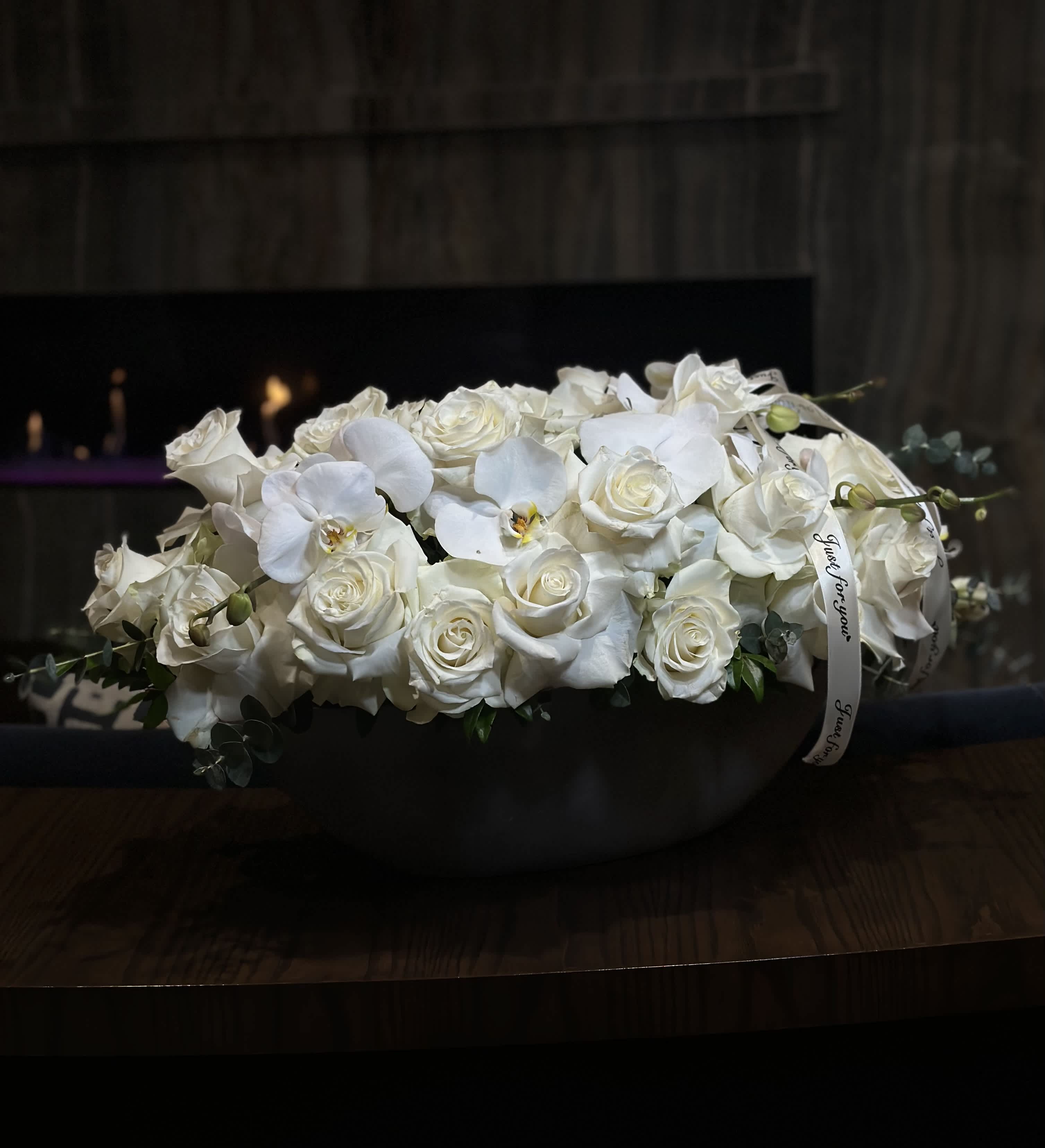White Swan - In a graceful gray clay vase, a scene of timeless elegance unfolds. White roses, their delicate petals unfurling like whispers of purity, stand tall and proud, exuding an aura of serenity and grace. Intertwined among them, slender branches of orchids extend gracefully, their exotic blooms adding a touch of mystique and sophistication to the composition. Against the subdued backdrop of the gray vase, the pristine white roses and ethereal orchids create a captivating contrast, evoking a sense of harmony and balance. Together, they form a breathtaking tableau, a celebration of natural beauty and refinement that enchants the eye and soothes the soul.