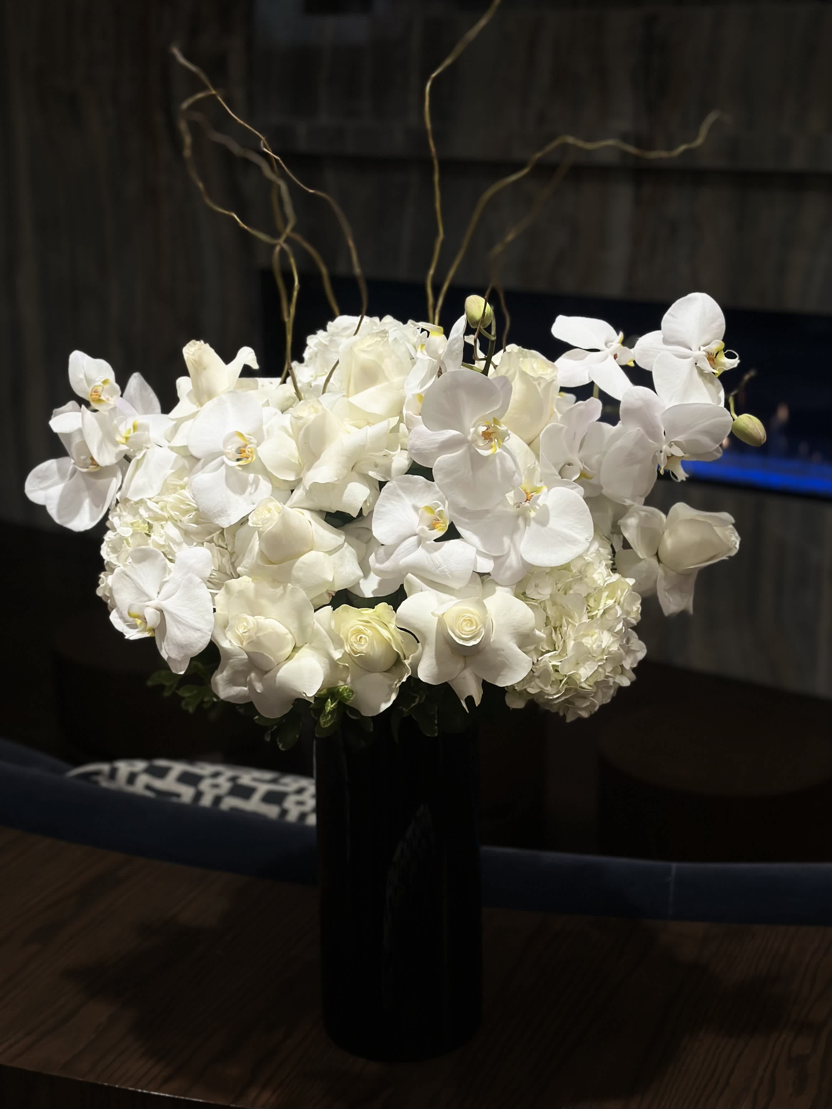 Black Pearl - A black vase arrangement with white orchids and roses is a striking blend of elegance and contrast. The sleek black vase provides a sophisticated backdrop for the pristine white blooms. The white orchids, with their delicate petals and graceful stems, exude a sense of purity and sophistication. Complemented by the timeless beauty of the white roses, the arrangement radiates a sense of refinement and luxury. This arrangement  is perfect for adding a touch of contemporary glamour to any space or event, evoking a sense of timeless beauty and sophistication.