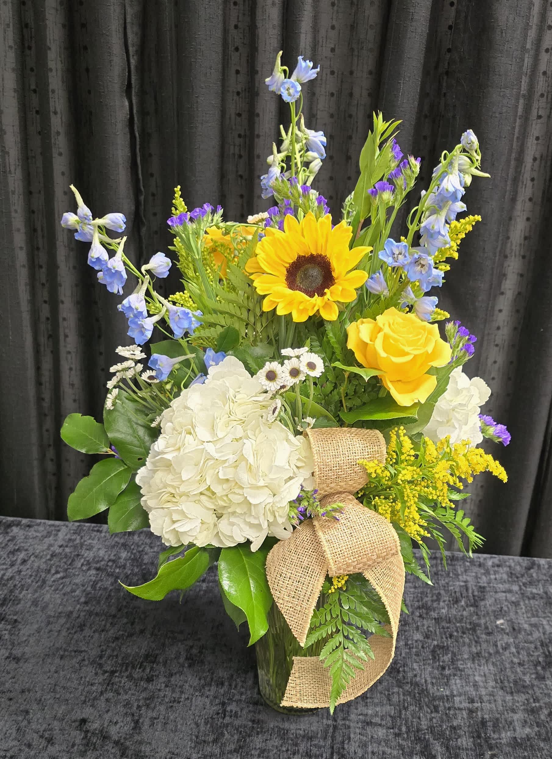 Country cottage. - A rustic arrangement made with hydrangeas, sunflowers, roses and delphinium and assorted filler flowers tied with a burlap ribbon