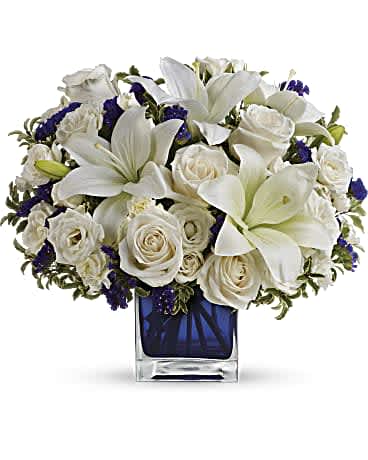 Sapphire Skies Bouquet - Like gazing into a clear blue sky, this serene arrangement soothes the soul and cheers the heart. Its creamy roses and snowy lilies are arranged in our dazzling sapphire cube. Crème roses, white asiatic lilies and white miniature carnations are mixed with bursts of purple statice and green