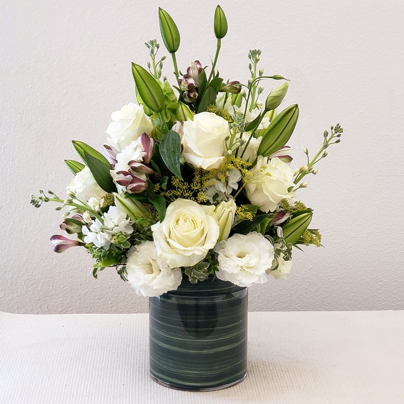 Eternal Blooms - Eternal Bloom of all-white seasonal flowers delivered in a cylinder glass vase. This bouquet exudes elegance with its pristine white roses and lush greenery  It's a timeless gesture perfect for any occasion, from heartfelt sympathies to sincere congratulations.  Approximately 17&quot; H x 15&quot; W. 
