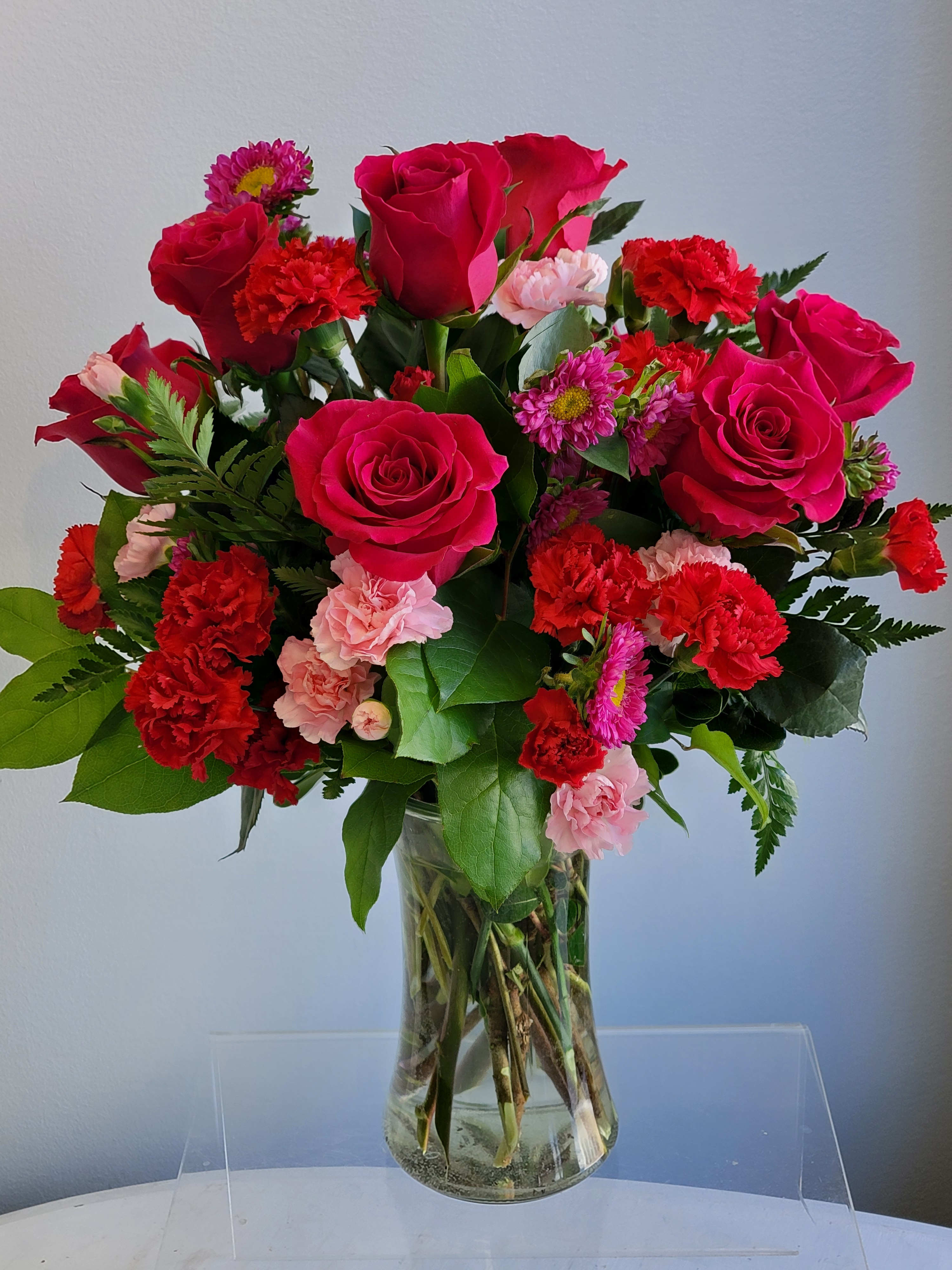 The Pink Floyd - Bright pink roses, Matsumoto asters, pink and red mini carnations arranged with mixed greens in a clear gathering vase