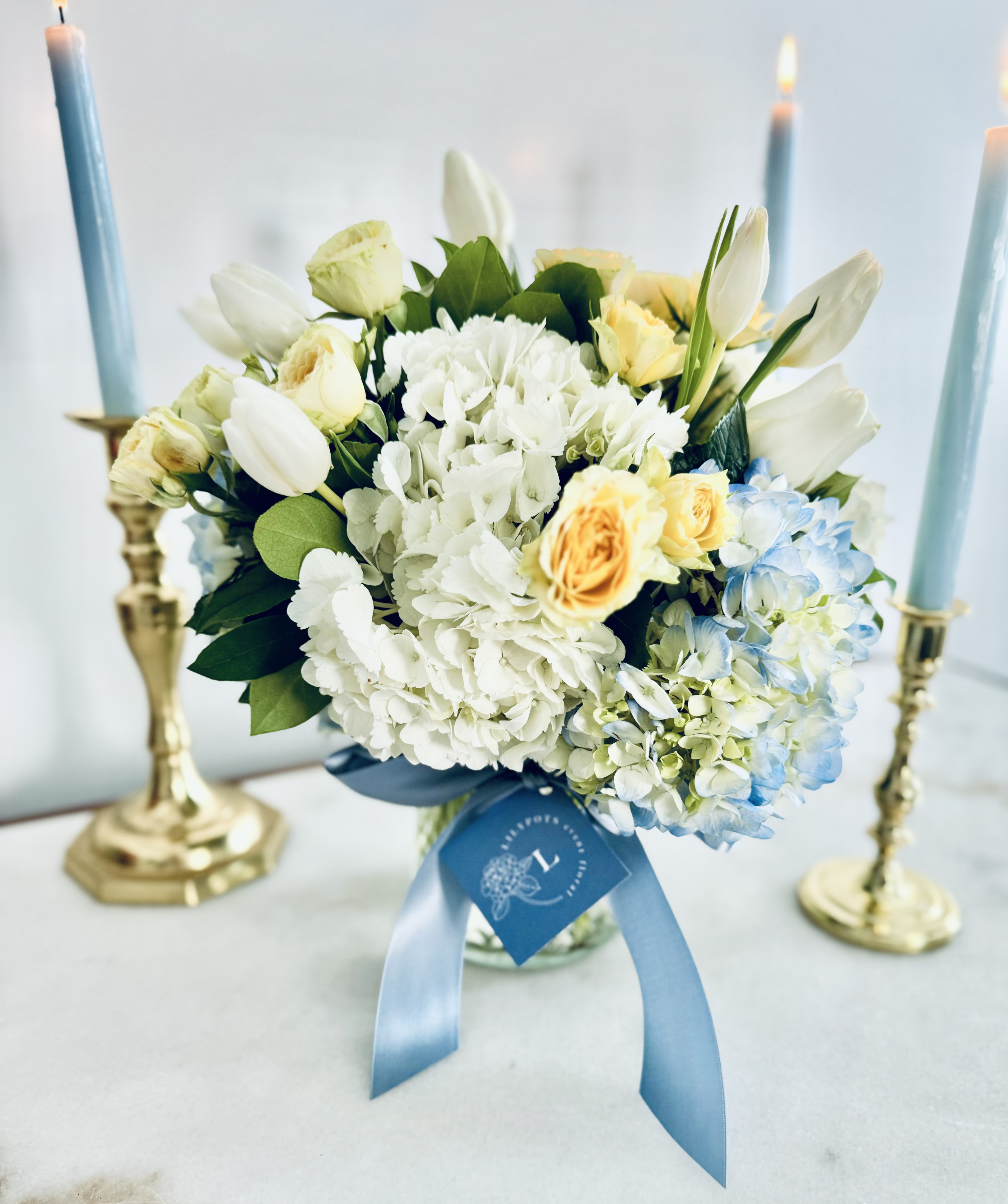 Geneva Lake Sunshine - A picturesque blend of yellow and blue blossoms reminiscent of a serene, sun-soaked day by Geneva Lake. The gentle sway of the petals echoes the tranquil ripples of the water's surface, while the vivid hues of the flowers reflect the bright radiance of the summer sun. This floral masterpiece perfectly captures the lively essence of the lake life.   CANDLES NOT INCLUDED  To maximize the longevity and aesthetic appeal of our flowers, we recommend refreshing the water in your arrangement daily. Additionally, it's advisable to keep your arrangement away from all heat sources, such as warm air drafts, direct sunlight from windows, and stoves, among others.  As flowers are a natural product, it may be necessary to make substitutions of floral varieties on occasion due to various factors. The substituted florals will be selected based on their value, color, and overall aesthetic to match the original arrangement shown in the photos.