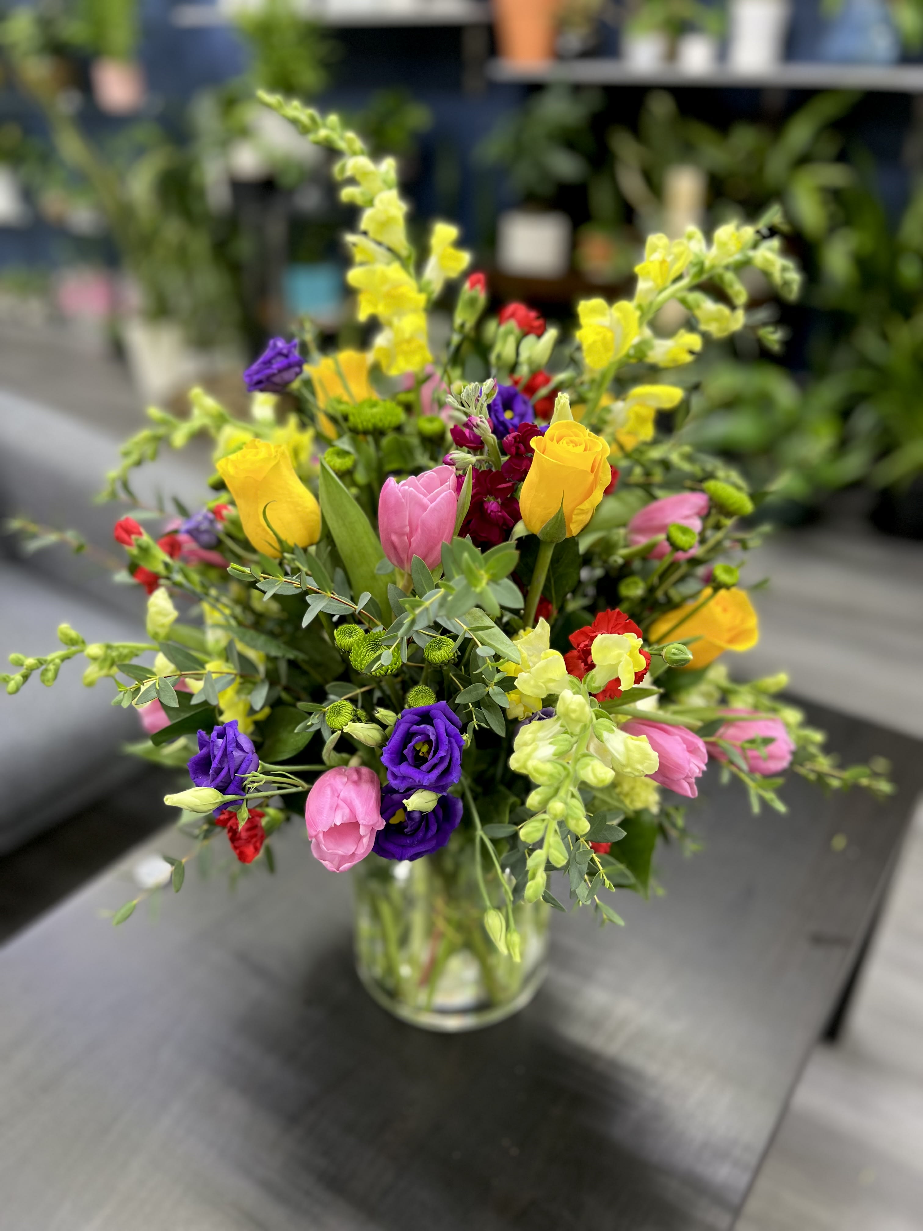 "Brushstrokes of Color in Height" "Brushstrokes of Color in Height" - "A majestic tall arrangement in an elegant glass vase, where vibrant strokes of color come together harmoniously. The tall snapdragons add verticality, while purple lisianthus, yellow roses, pink tulips, burgundy stock, and red carnations intertwine to create a unique floral symphony. This arrangement is a visual feast that celebrates the diversity of colors and elegance, perfect for brightening any special occasion with its striking presence and floral charm."
