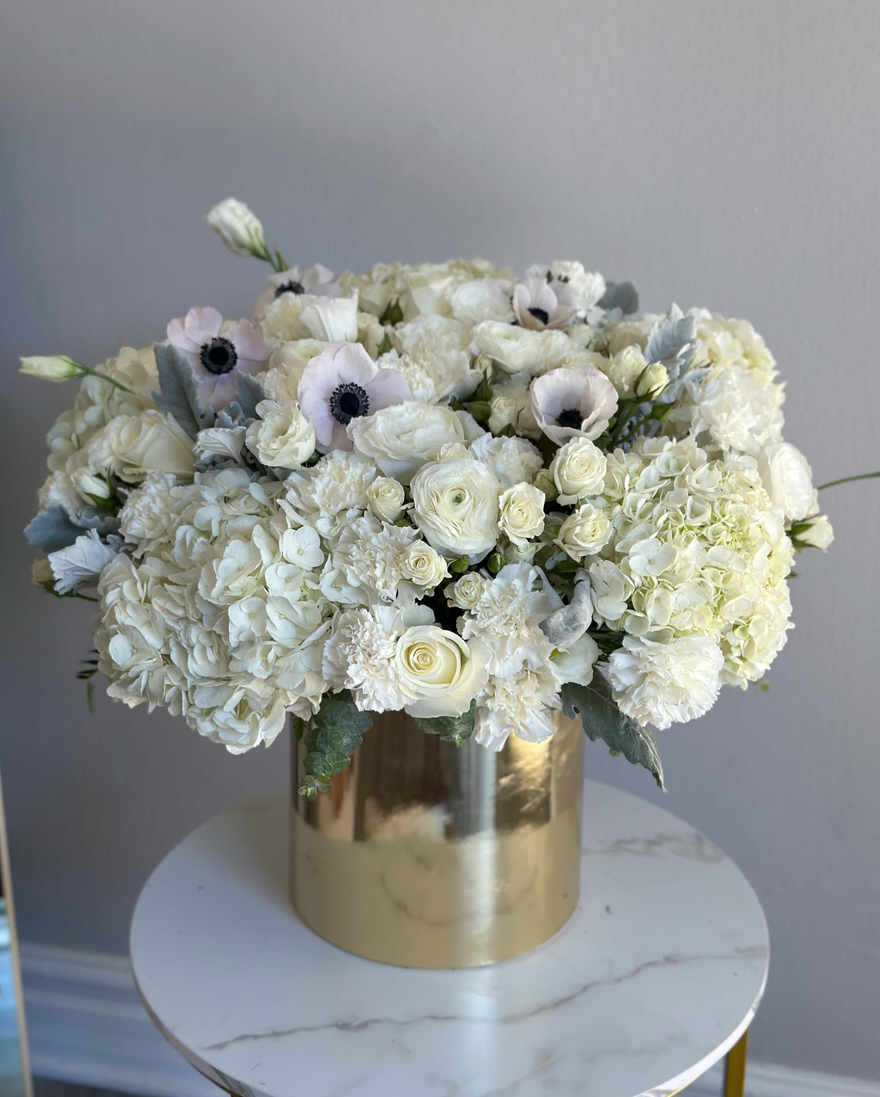 Golden Box with White Anemones - Exquisite arrangement in a golden box, adorned with delicate white anemones, graceful roses, and lush hydrangeas. The white anemones add an air of mysterious charm to the composition, their elegant petals seeming to dance in the breeze. Roses, symbolizing love and beauty, infuse the atmosphere with romance and sophistication. Meanwhile, hydrangeas with their abundant clusters create an ambiance of abundance and refined allure. Together, these flowers form a magnificent combination that inevitably draws admiring glances, creating an atmosphere of refined beauty and timeless elegance. 