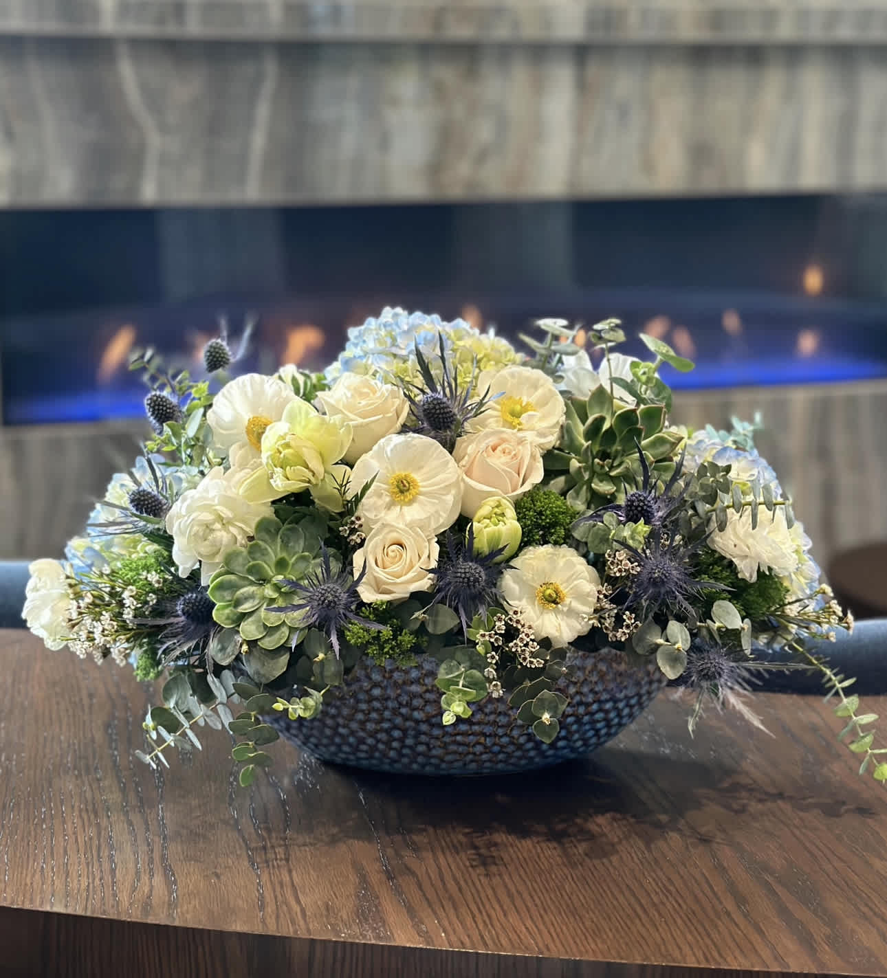 Through Thorns to the Stars  - In a rustic vase, a botanical masterpiece takes shape. Lush succulents, with their intricate patterns and vibrant hues, form the foundation of this enchanting arrangement, their hardy beauty adding a touch of earthy elegance. Nestled among them, hydrangea blooms unfurl in shades of blue and purple, their delicate petals invoking a sense of tranquility and grace. Adorning the composition, stems of eryngium add a striking contrast with their spiky texture and silver-blue hue, lending an air of wild beauty. Finally, ranunculus blooms burst forth in a symphony of color, their layers of petals unfolding like delicate satin. Together, they create a captivating tableau, a celebration of nature's diversity and resilience that captivates the eye and nourishes the soul. 