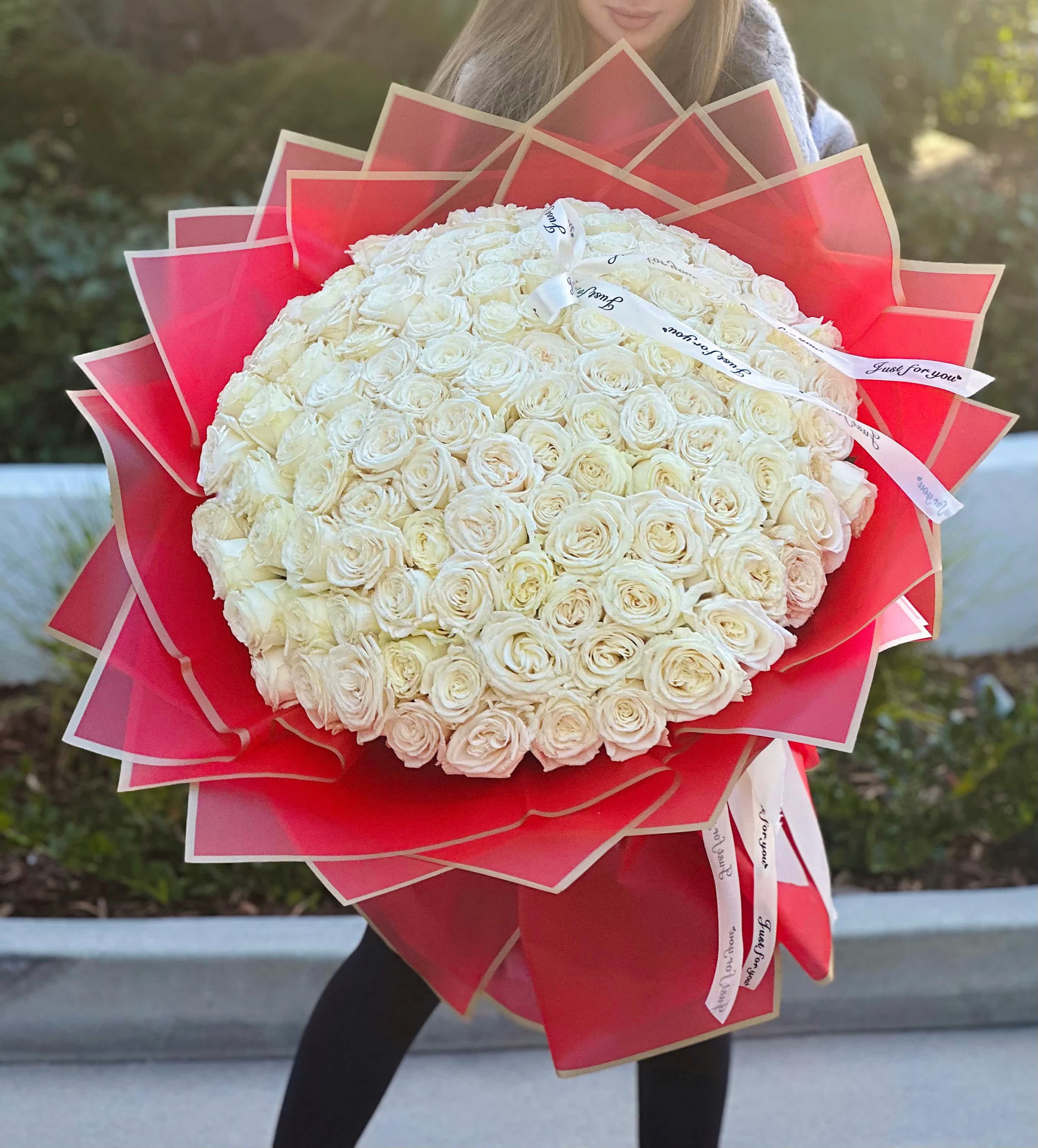 White Queens Bouquet  - A bouquet of 100 pristine white roses, cradled in vibrant red wrapping adorned with a golden trim, and adorned with elegant white ribbons, embodies the essence of purity and opulence. Each rose, like a symbol of innocence, radiates timeless beauty, exuding grace and elegance. The red packaging, accented with golden details, adds a touch of regal splendor, while the white ribbons dance delicately around the blooms, adding an air of sophistication to the ensemble. Together, they create a captivating tableau, a harmonious blend of purity and luxury that captivates the eye and warms the heart.