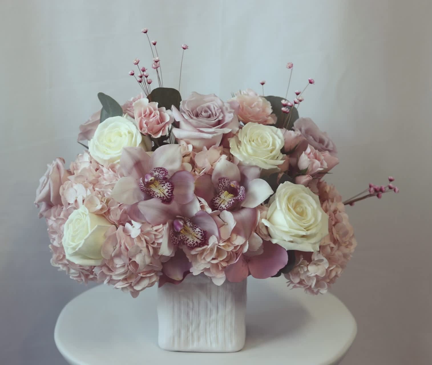 Pink Meadows Pink Meadows - Venetian Flowers has created a stunning bouquet for Mother's Day. This modern arrangement features a beautiful white ceramic cube linen print vase. The bouquet itself is a combination of elegant and delicate flowers. It includes pink hydrangea, pink cymbidium orchids, white Playa Blanca roses, and dusty mauve pink roses. The bouquet is complemented by filler flowers and eucalyptus, adding texture and a touch of greenery. The overall design is contemporary and sophisticated, perfect for a modern aesthetic. The high-end cube linen print vase adds an extra element of luxury to this stunning arrangement. Standard- 3 pink large hydrangeas, 9 premium roses, 3 large cymbidium orchids, fillers and eucalyptus. Deluxe - 4 large hydrangeas, 12 roses, 4 orchids, euc and filler flowers in a slightly larger 5x5 linen print ceramic cube. Premium - 5 large hydrangeas, 15 roses, 5 large orchids, euc and fillers in a 5x5 linen print modern modern ceramic cube vase.