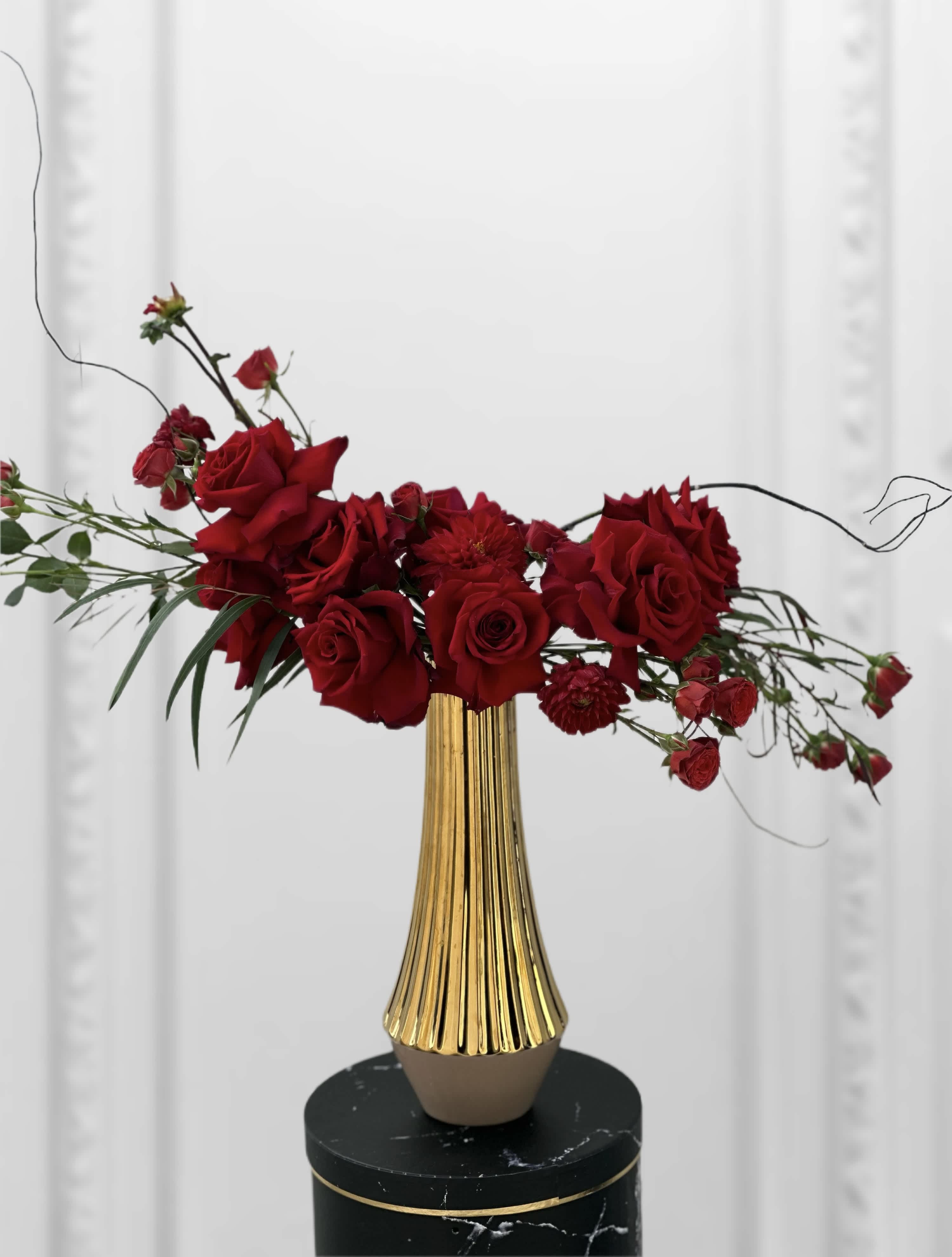 Golden Nuance Roses  - In a majestic tall golden vase, a scene of regal elegance unfolds. Crimson roses, their velvety petals unfurling like whispers of passion, stand tall and proud, exuding timeless beauty and romance. Intertwined among them, slender branches extend gracefully, their delicate foliage adding a touch of natural allure and movement to the composition. Against the gleaming backdrop of gold, the rich red hues of the roses radiate with a mesmerizing allure, creating a captivating tableau that commands attention and ignites the imagination.