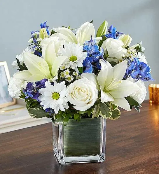 Healing Tears  Blue &amp; White - Soothe their tears as they mourn the loss of a loved one with the serenity of blue and white. Our sympathy arrangement of fresh blue delphinium, white roses and lilies, expertly gathered together in a clear glass cube lined with Ti leaf ribbon, makes for an exquisite gesture of comfort and healing.