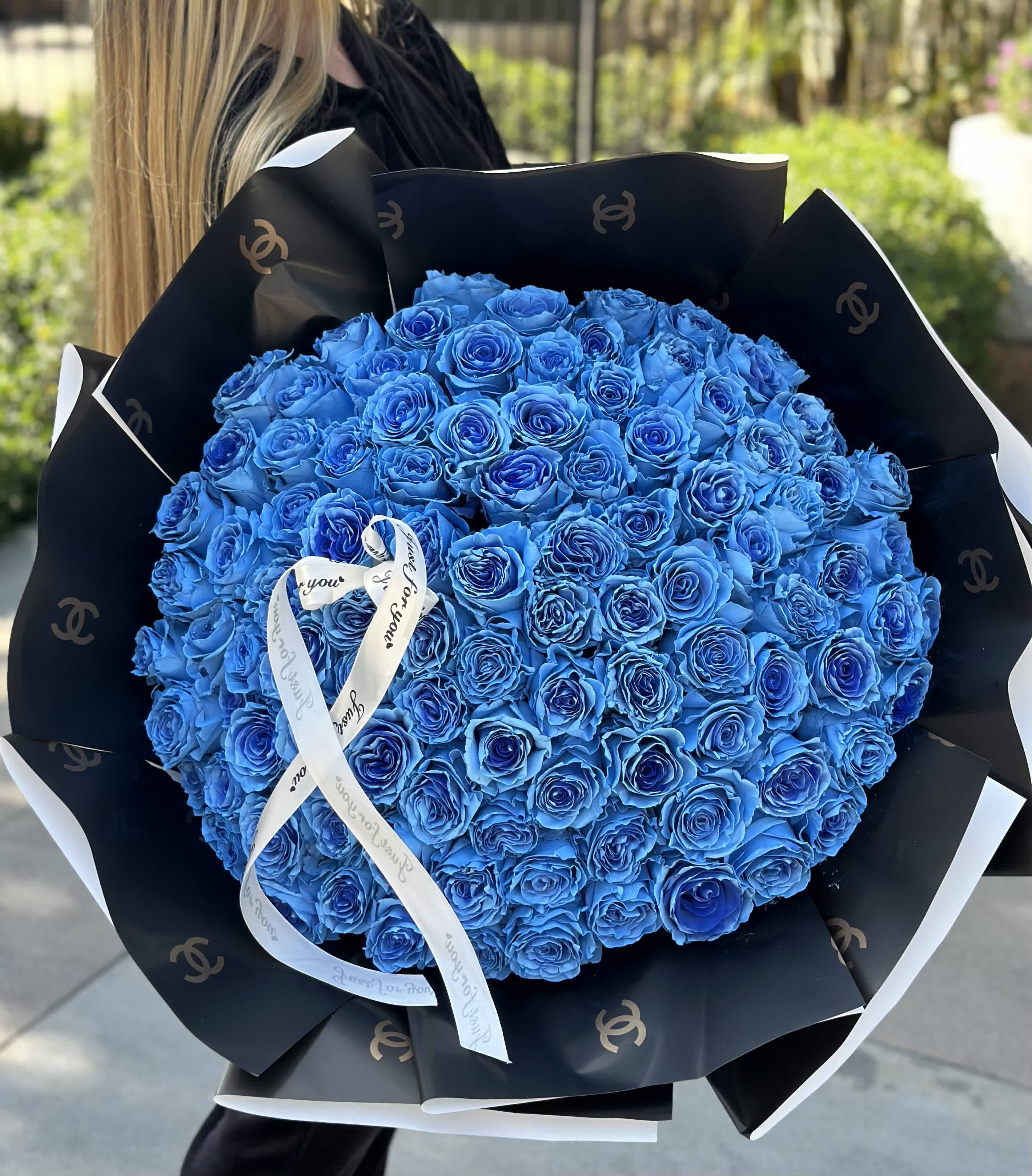 100 Blue Roses Wrapped - A bouquet of azure roses nestled within sleek black packaging, adorned with flowing ribbons, creates a striking visual symphony. Each bloom, reminiscent of the vast expanse of the sky, emanates tranquility and depth, capturing the essence of serenity. The black packaging serves as a dramatic backdrop, enhancing the vividness of the blue blooms and adding an element of mystery to the arrangement. Delicate ribbons dance around the bouquet, adding a touch of elegance and movement, completing the ensemble with a flourish of grace. Together, they compose an enchanting tableau, a fusion of boldness and refinement that captivates the senses and evokes a sense of wonder.