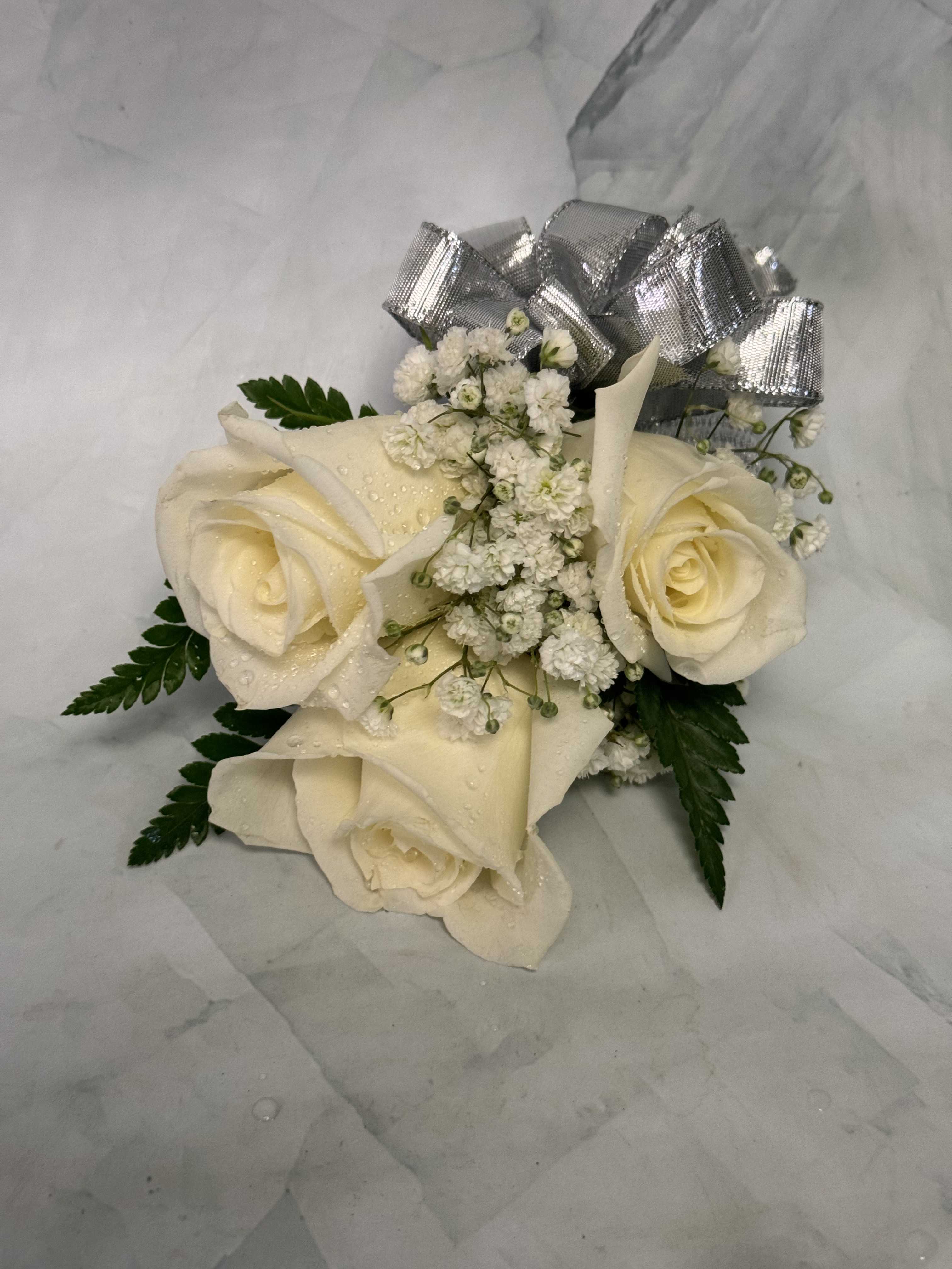 Traditional White Rose and Silver Bow Wrist Corsage - This is a traditional take on a wrist corsage. Each one of our corsages is custom and hand-made. This corsage includes three roses, greenery, babies breath and a silver bow. 