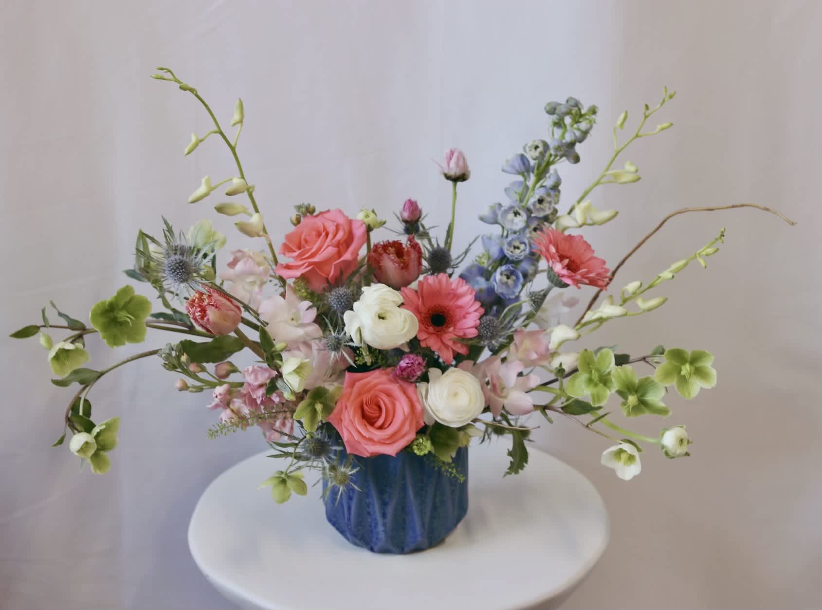 Blue Splendor  - Venetian Flowers has created the Blue Splendor - a vibrant mixed bouquet that is sure to make a statement. This stunning arrangement is presented in a modern blue geometric vase, adding a contemporary touch to the overall design. The bouquet features a variety of beautiful flowers, including orchids, gerbera daisies, tulips, delphinium, hellebores, and ranunculus. The combination of these flowers creates a colorful and eye-catching display. To add an extra touch of elegance, the bouquet is accented with premium greens and curly willow, adding texture and dimension. The arrangement is a perfect blend of modern style and natural beauty, making it a fantastic choice for any occasion.