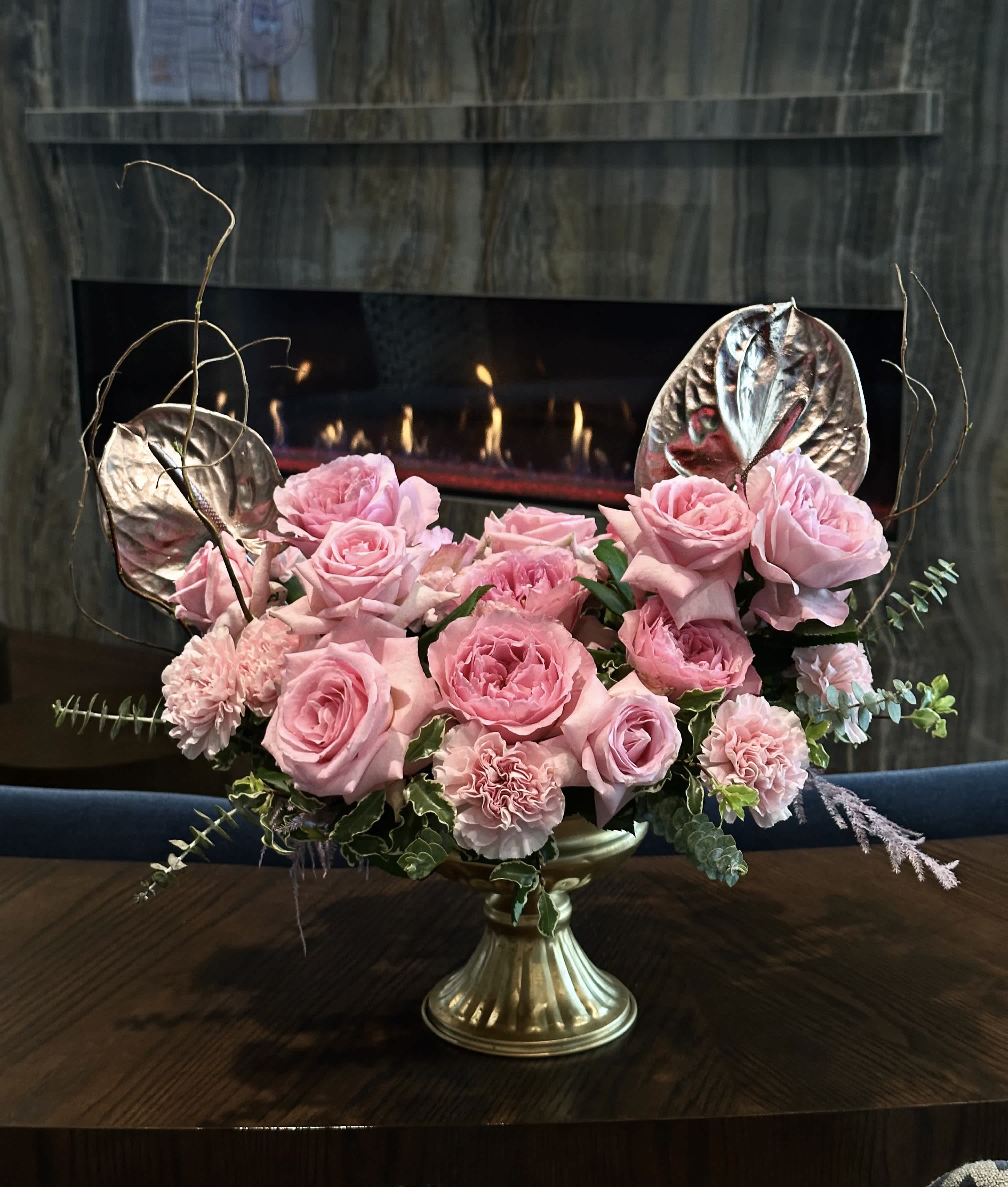 Floral euphoria - In a golden vase, a mesmerizing arrangement unfolds, embodying elegance and grace. Delicate branches weave through the composition, adding a touch of natural whimsy. Soft, blush-pink roses bloom with timeless beauty, their petals gently unfurling. Amidst them, striking anthuriums stand tall, their vibrant hues and unique shapes offering a captivating contrast. This pink ensemble in its lustrous golden vessel evokes a sense of refined luxury and delicate charm, making it a perfect centerpiece for any setting.