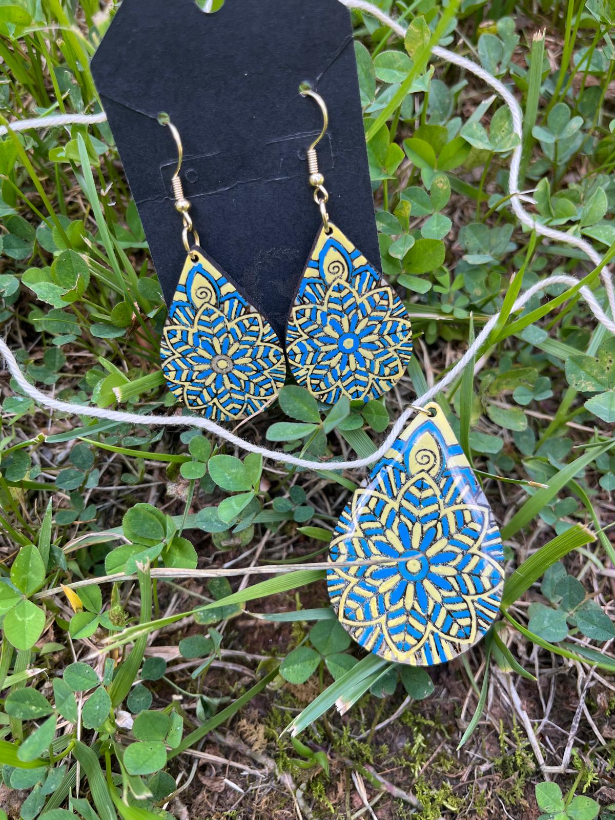 Slava Ukraine Flower Mandala Earrings - Show your support for Ukraine with this stunning Slava Ukraine Flower Mandala jewelry set. 10% of proceeds for each sale support Ukrainian Refugees.   Handcrafted with love by Virginia artisans, this unique set features intricate laser-cut wood designs inspired by traditional Ukrainian flower mandalas. Each piece is meticulously hand-painted and sealed with resin for a vibrant, long-lasting finish.  Key Features:  Unique Design: Each piece showcases a beautiful and intricate floral mandala design, symbolizing Ukrainian culture and resilience. Handcrafted Quality: Made with care by skilled Virginia artisans, ensuring exceptional craftsmanship and attention to detail. Vibrant Colors: Hand-painted with vibrant colors that capture the spirit of Ukraine and the beauty of its floral motifs. Durable Construction: Laser-cut wood construction sealed with resin for long-lasting wear and a glossy finish. Complete Set: Includes a pair of earrings and a necklace pendant on a gold-plated chain (chain not pictured), perfect for layering or wearing separately. Meaningful Gift: A thoughtful and meaningful gift for anyone who supports Ukraine or appreciates unique, handcrafted jewelry. Additional Details:  Materials: Laser-cut wood, resin, paint, gold-plated chain  Wear this beautiful jewelry set as a symbol of solidarity with Ukraine and a celebration of its rich cultural heritage.  Note:  Chain not pictured, but included with purchase.