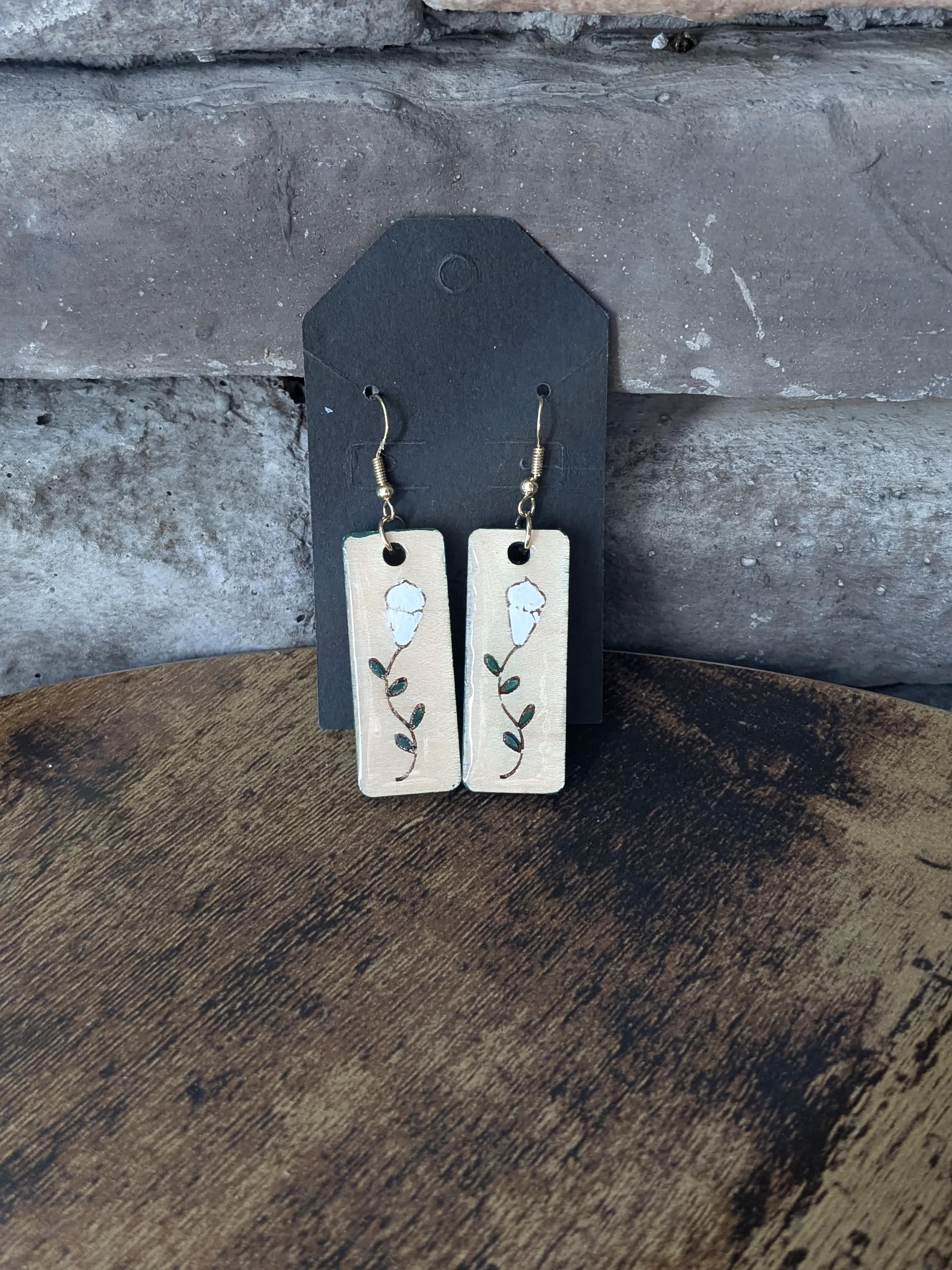 Virginia Lily Lace Earrings - Handcrafted Maple Wood Earrings: Virginia Lily Lace  Adorn your ears with the delicate elegance of these unique earrings, meticulously handcrafted by Virginia artisans. Each pair features smooth maple wood discs, carefully laser-engraved with intricate lily designs that capture the essence of nature's purity and grace.  The engraved lilies are then lovingly hand-painted in luminous white acrylics, showcasing the artistic skill and individual touch of the artisans. A protective layer of resin is applied to seal and enhance the delicate details, ensuring their lasting beauty.  No two pairs are exactly alike, making these earrings a truly special and personal accessory. Embrace the one-of-a-kind charm of these handcrafted treasures and let the ethereal beauty and intricate details elevate your everyday style.  Features:  Materials: Maple wood, acrylic paint, resin Technique: Laser engraving, hand painting Origin: Handcrafted in Virginia, USA Style: Unique, nature-inspired