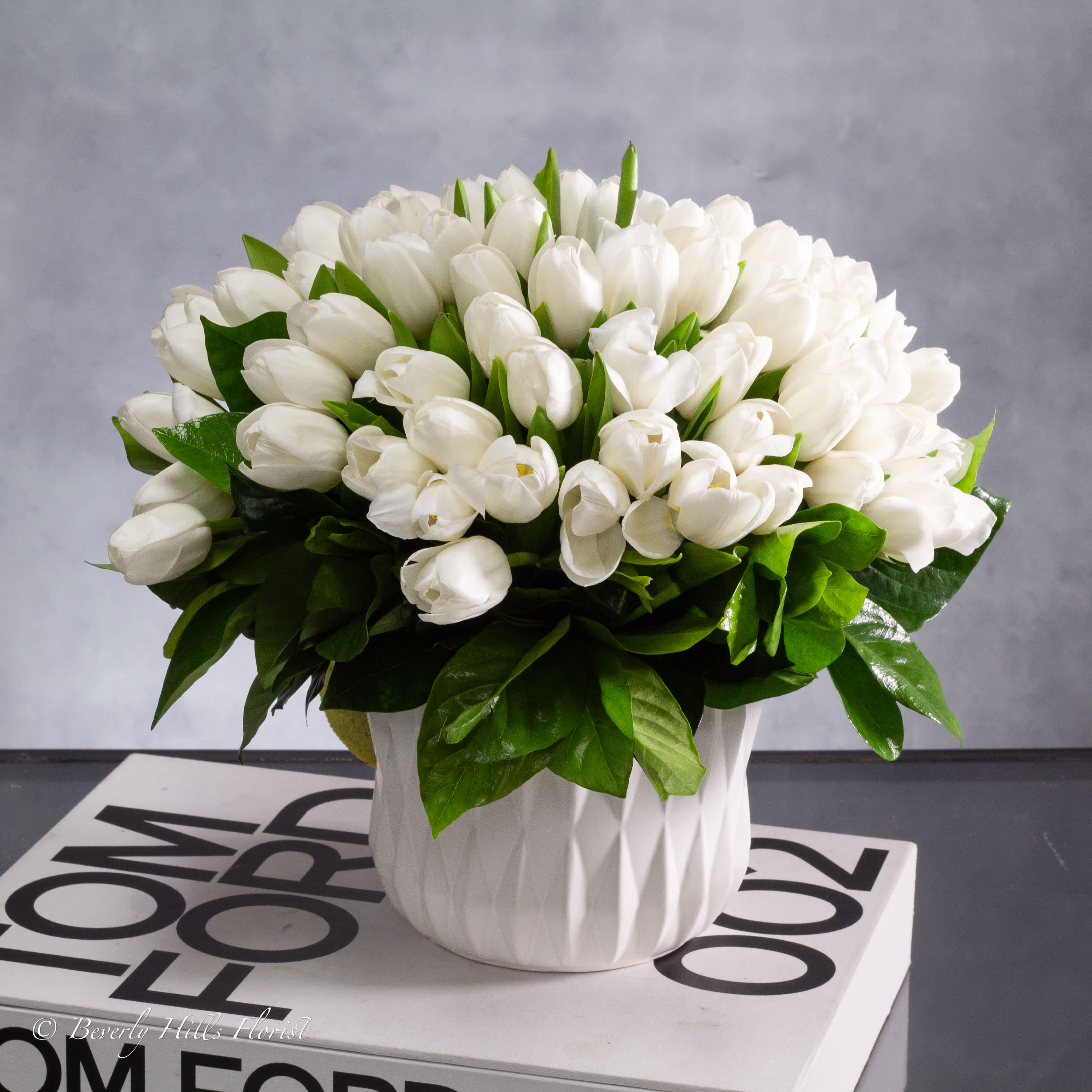 White Tulips On White - Simple elegance at it’s best!  A lovely bouquet of about 80 white Tulips stand atop beautifully arranged picturesque Modern Vase.  A beautiful expression of your sweetest sentiments!  Measurement   10&quot; Tall and 10&quot; Wide in a 5&quot; Geometric Vase