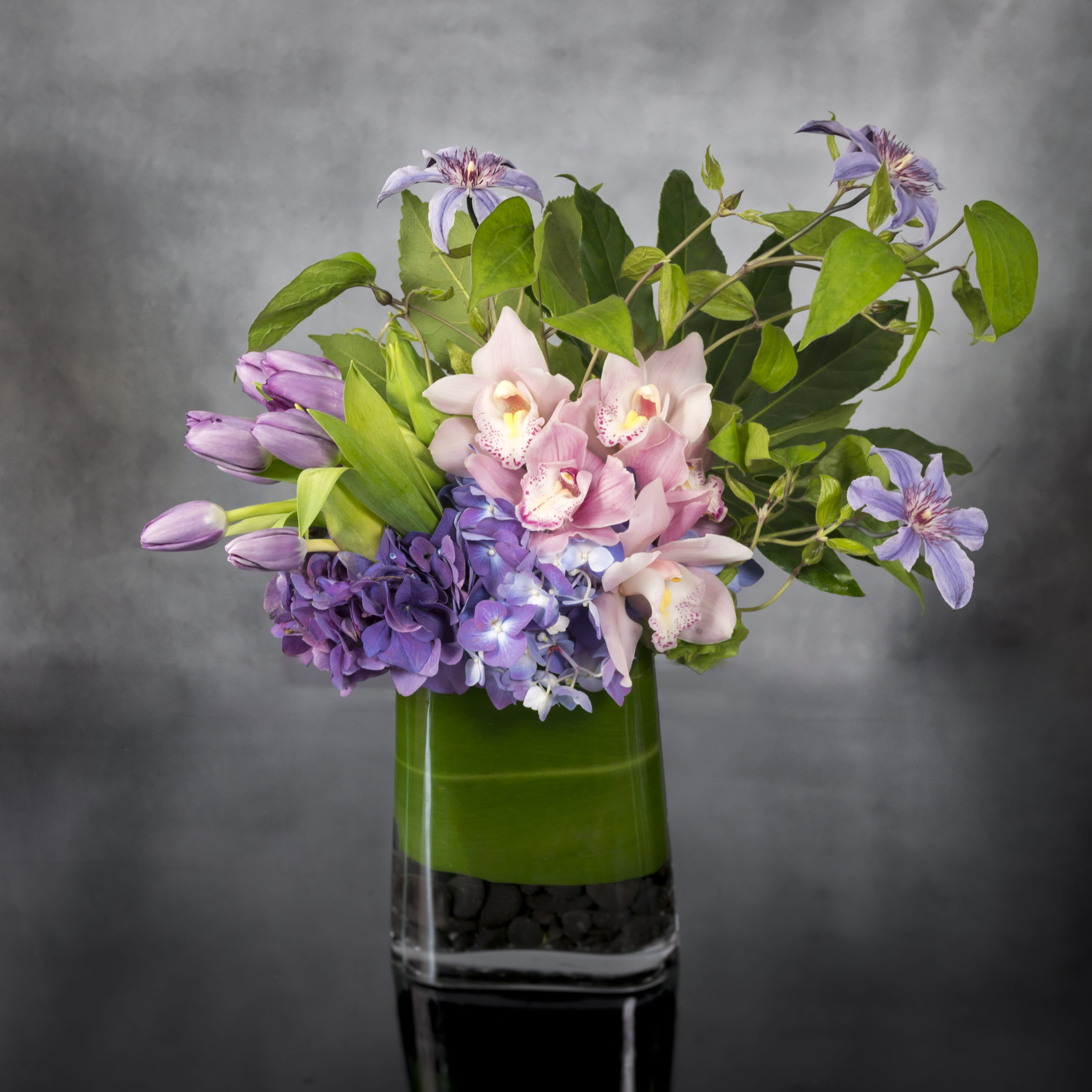 Just the Way You Are - Express your love with this beautiful floral collage of lavender Tulips, pink cymbidium Orchids, purple Hydrangeas and more a classic leaf-lined glass vase filled with river rocks.    Standard:  Arrangement Approx. 12&quot;-14&quot; Tall  in a 7&quot; Rectangular Container 
