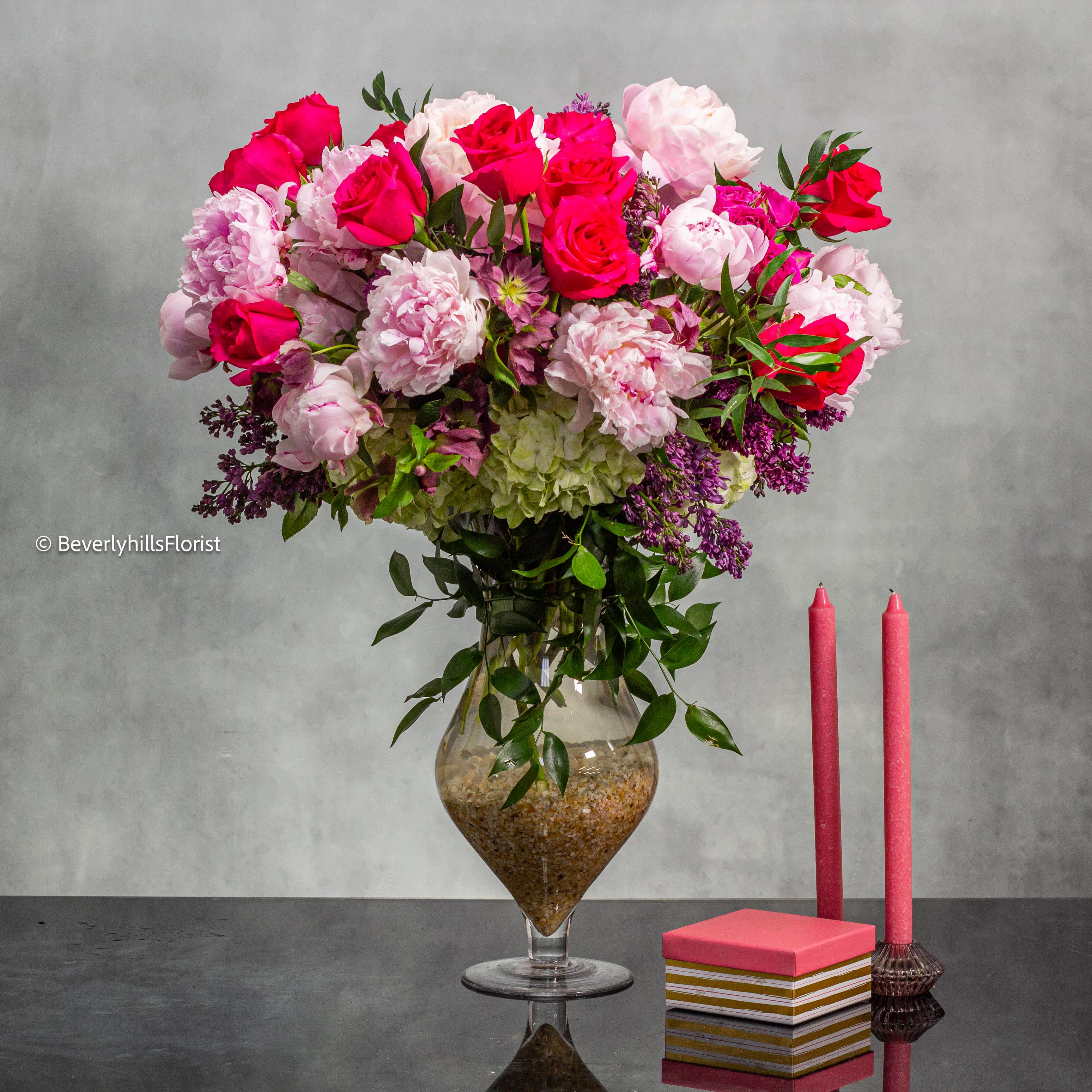 Luxe Blooms of Milan and Netherlands - Elevate your surroundings with this opulent floral arrangement that marries the finest blooms from Milan and the Netherlands. Standing at approximately 28 inches tall and 24 inches wide, this masterpiece features a captivating blend of hydrangeas, lilacs, hot pink roses, spray roses, and fresh Italian peonies that have been imported directly from Milan for an unrivaled touch of luxury.  Adorned in a beautiful orange vase, this arrangement exudes sophistication and elegance, making it a perfect statement piece for any discerning recipient in Beverly Hills and surrounding areas. Each bloom, meticulously selected for its beauty and quality, harmonizes to create a symphony of colors and fragrances that will enchant the senses. With same-day delivery available, indulge in the lavish beauty of these exquisite blooms delivered straight to your doorstep.