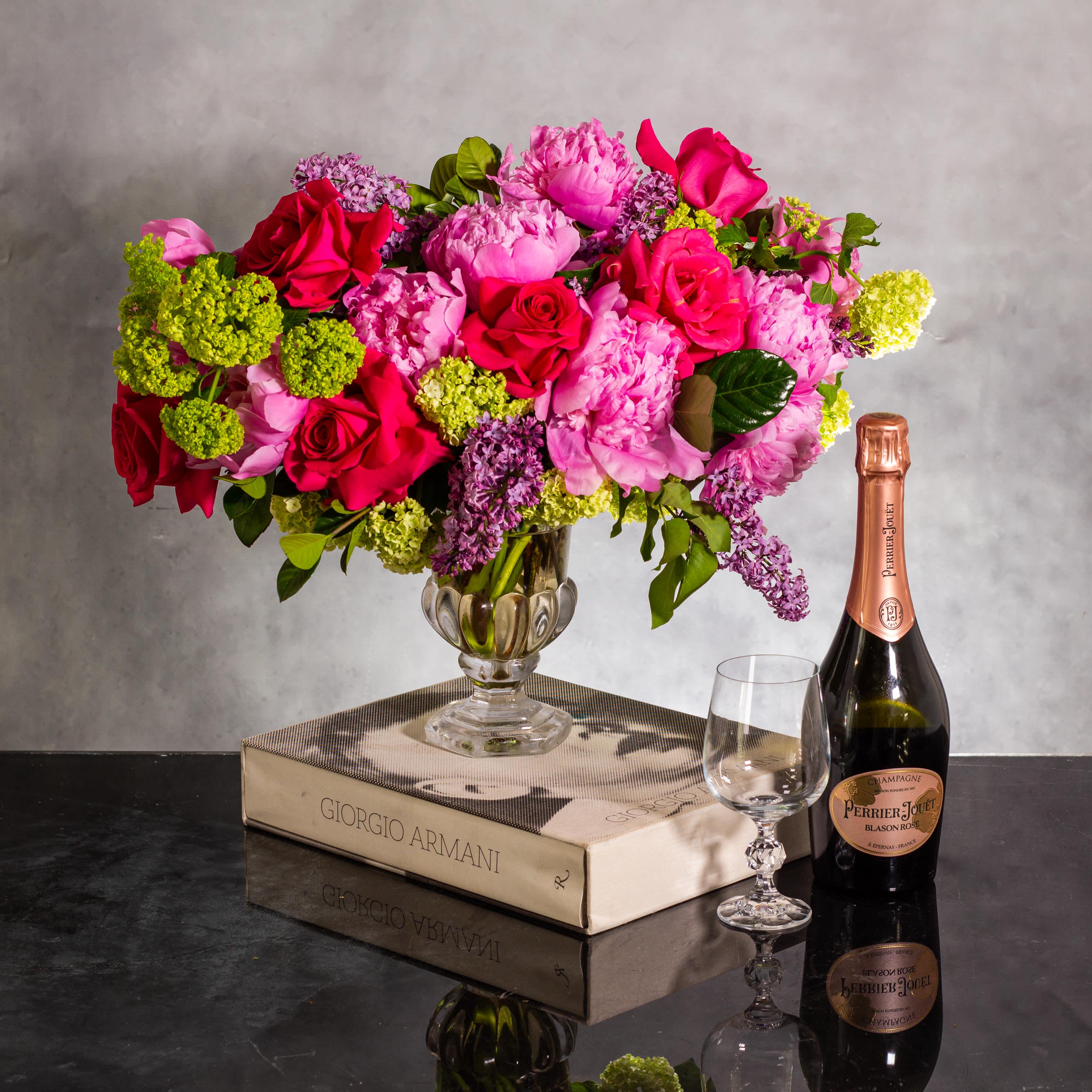 Lilac Petals and Peonies - Secrets of the Viburnum - The floral arrangement is a sight to behold, showcasing large-headed pink peonies alongside hand-opened hot pink roses, viburnum, and lush, beautiful foliage. Placed in a pedestal glass vase standing approximately 14 inches tall and 16 inches wide, the arrangement adorns a 7-inch crystal vase. The combination of textures and colors in this arrangement must create a truly captivating and elegant centerpiece.