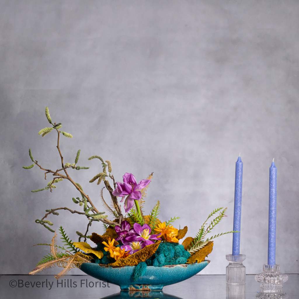 WILD WHISPERS - AN ENCHANTED GARDEN - Imagine a captivating scene where the untamed beauty of nature meets the elegant touch of artistic design. Our wild arrangement, nestled in an oval dish, features branches tinted moss to evoke the allure of a lush forest glade. Within this enchanting setting, our talented floral designers artfully arrange a stunning array of tulips in a myriad of colors, shapes, and sizes. Each tulip is carefully selected as part of our Designer's Choice, ensuring a unique and vibrant composition that reflects the essence of nature's bounty. Let this enchanting oasis bring a touch of wilderness and whimsy into your space, creating a mesmerizing focal point that captivates the imagination. 15&quot; Long ceramic container