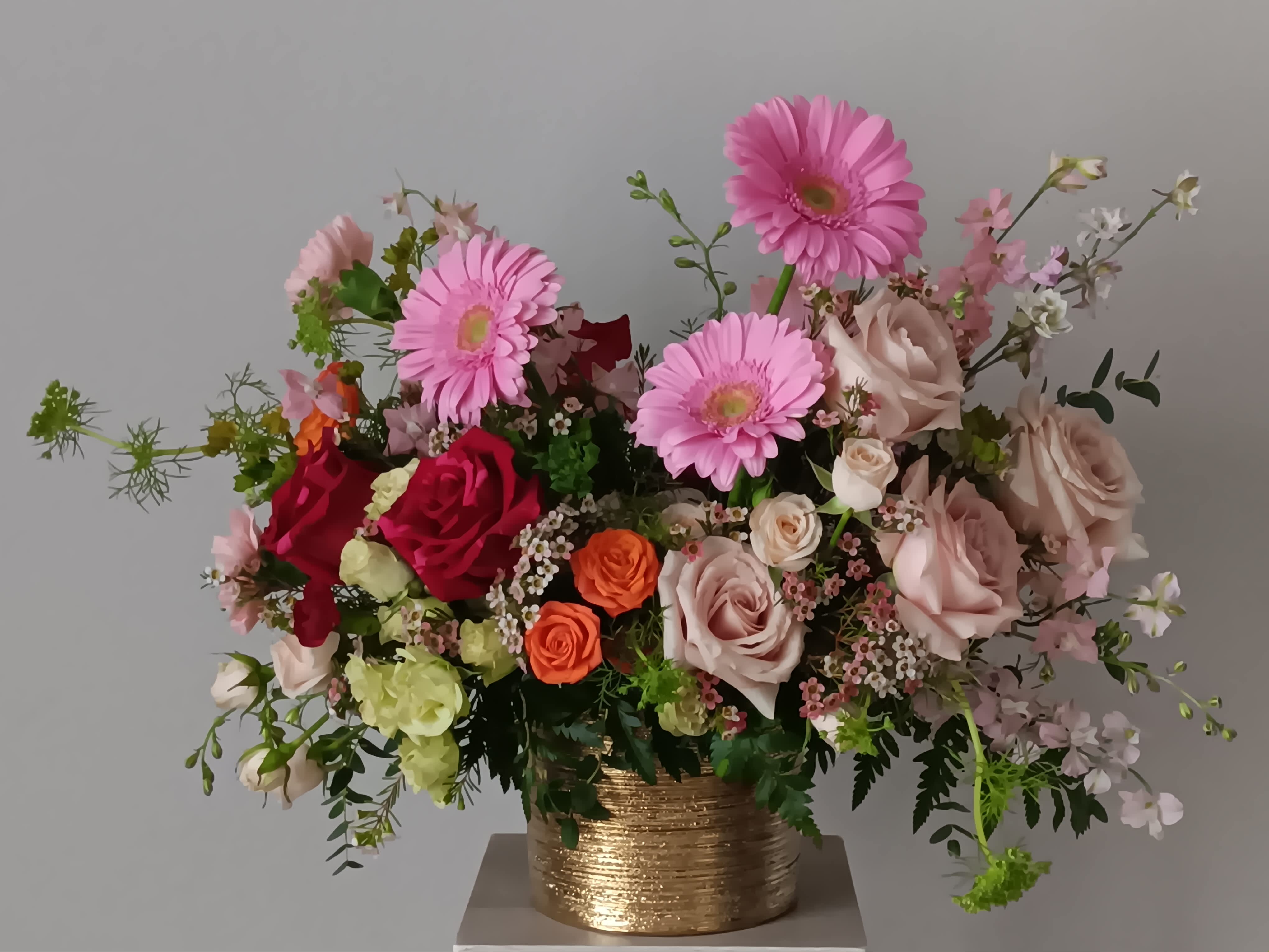 Birthday Brights Bouquet - The Birthday Brights Bouquet is a true celebration of color and life to surprise and delight your special recipient on their big day! Pink gerbera daisies and orange roses take center stage surrounded by red and light pink roses, spray roses, carnations, wax flowers, and lush greens to create party perfect birthday display. Presented in a modern gold ceramic vase. Orientation: All-around.   SUBSTITUTION POLICY – Always deliver the freshest flowers! Please note the bouquet pictured reflects our original design.  If the exact flowers or container in this arrangement are not available, our local florists will create a beautiful bouquet with the freshest available flowers.