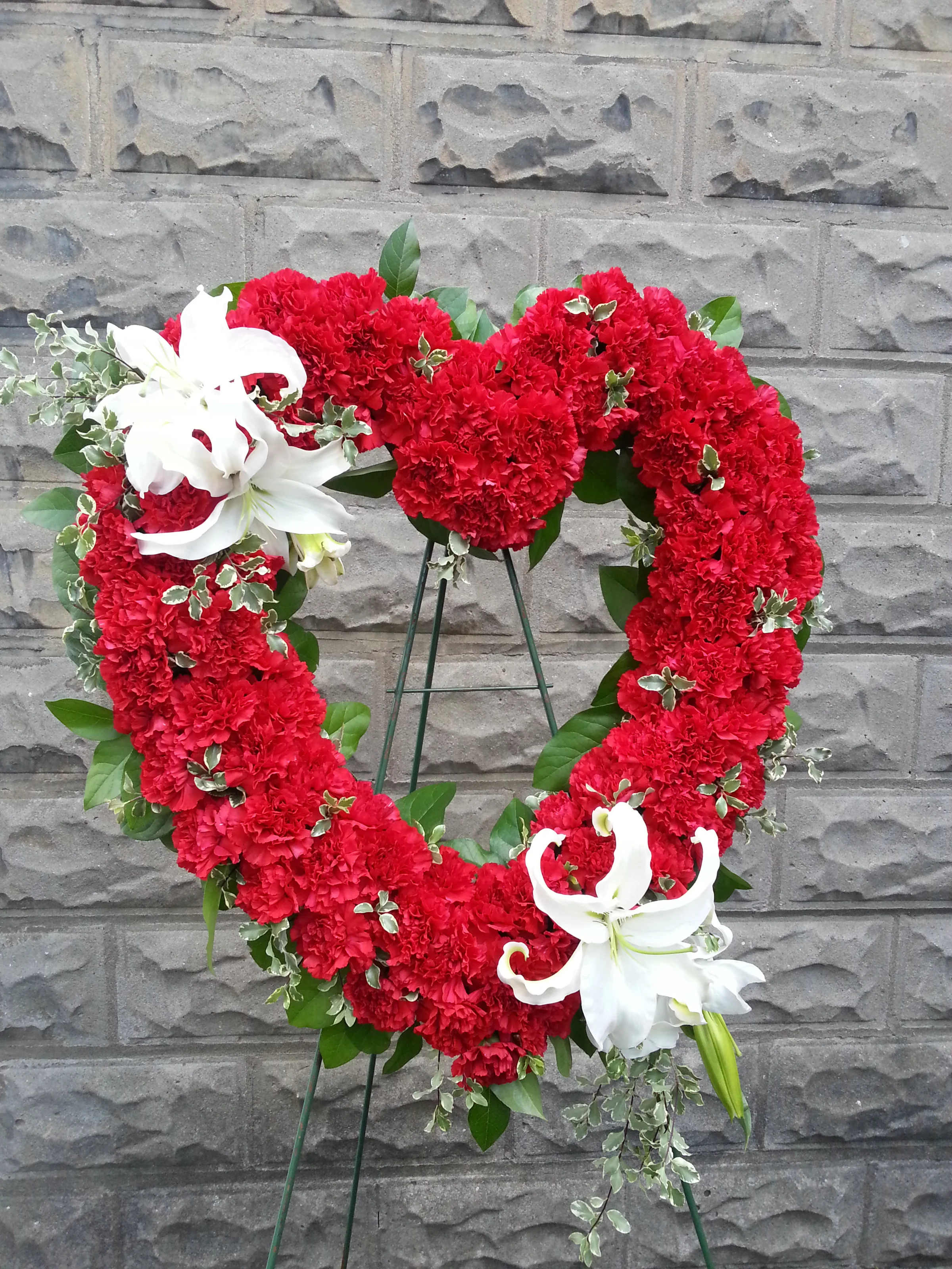 Solid Red Wreath with Lilies Solid Red Wreath with Lilies - These wreaths are vibrant expression of love. These can be done in an open heart or a wreath shape and can be created in a variety of colors. Please Note: Each design is custom made and filled to value. We only use fresh product to ensure top quality so new orders are subject to substitutions.